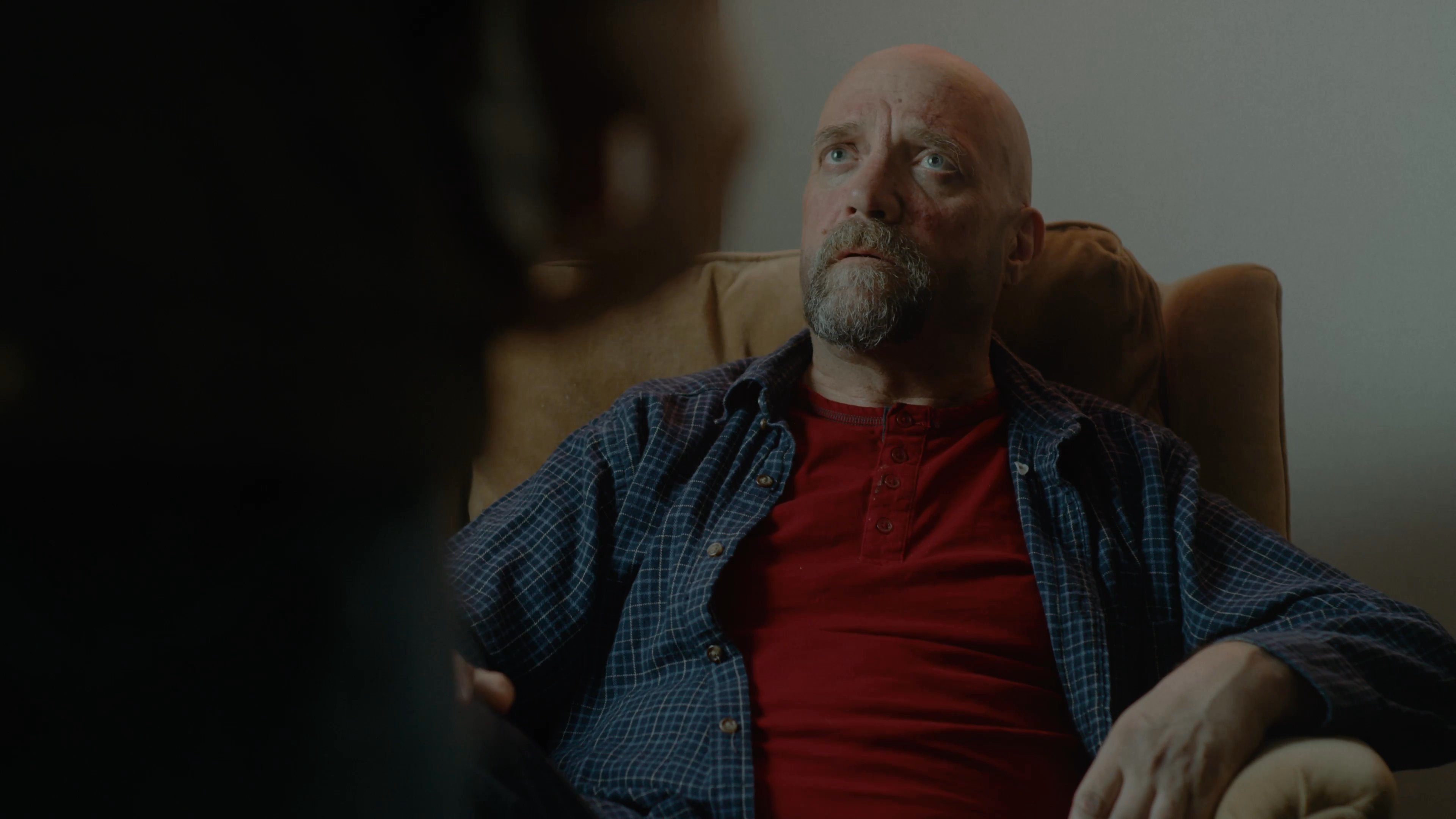 Two men engage in a calm, seated conversation within a casual home living space decorated with sports memorabilia.