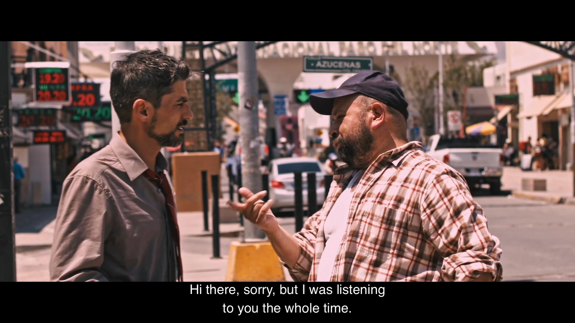 Two men standing on a city street discuss their shared experience of being deported. They engage in a somber conversation about the current challenges they face.