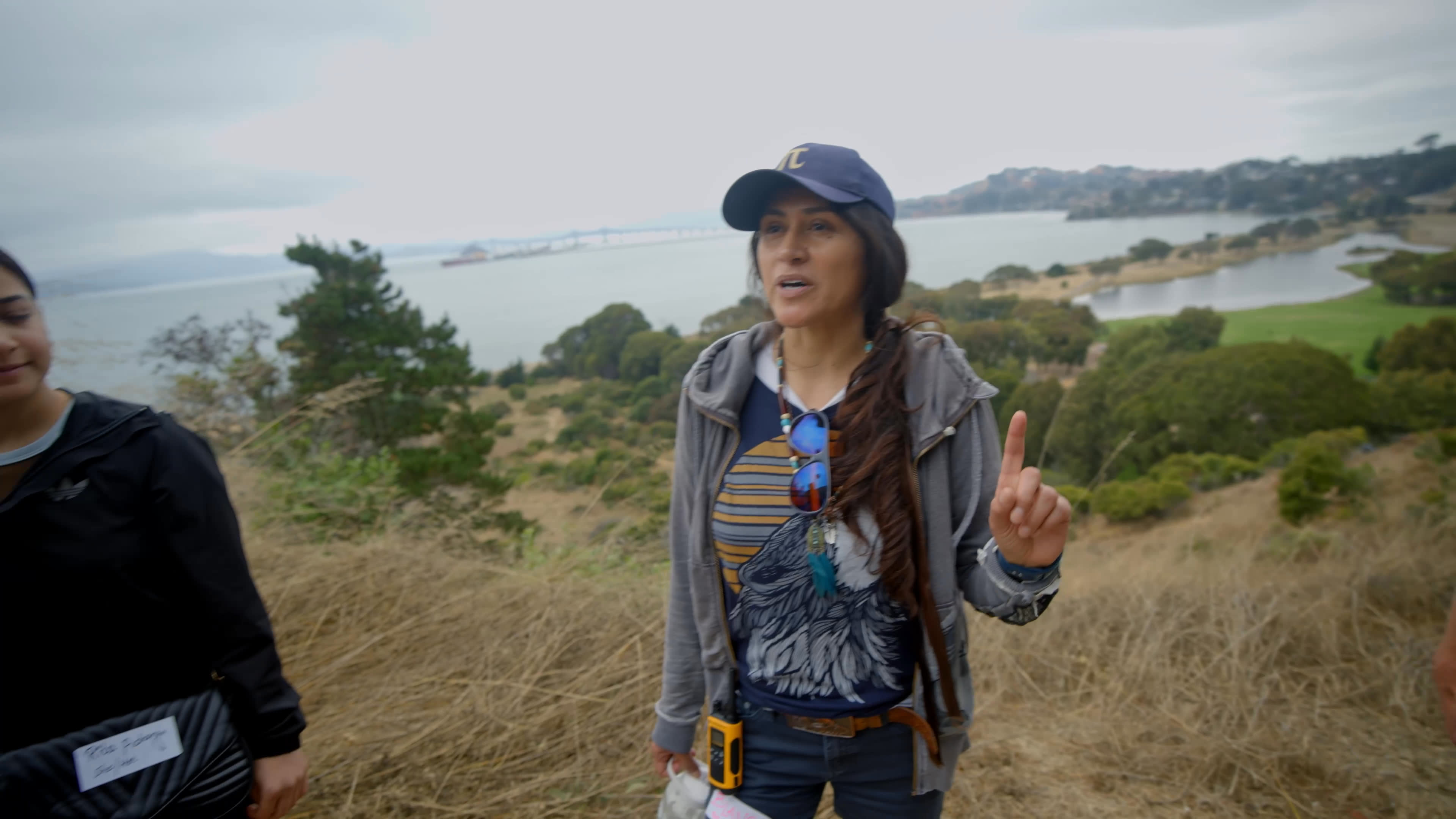 A guide gathers a group on a dry coastal hillside and asks who lived on the land before the refinery, train tracks, and bridges. The group looks across the bay toward the industrial shoreline as she invites them to imagine the landscape without those structures.