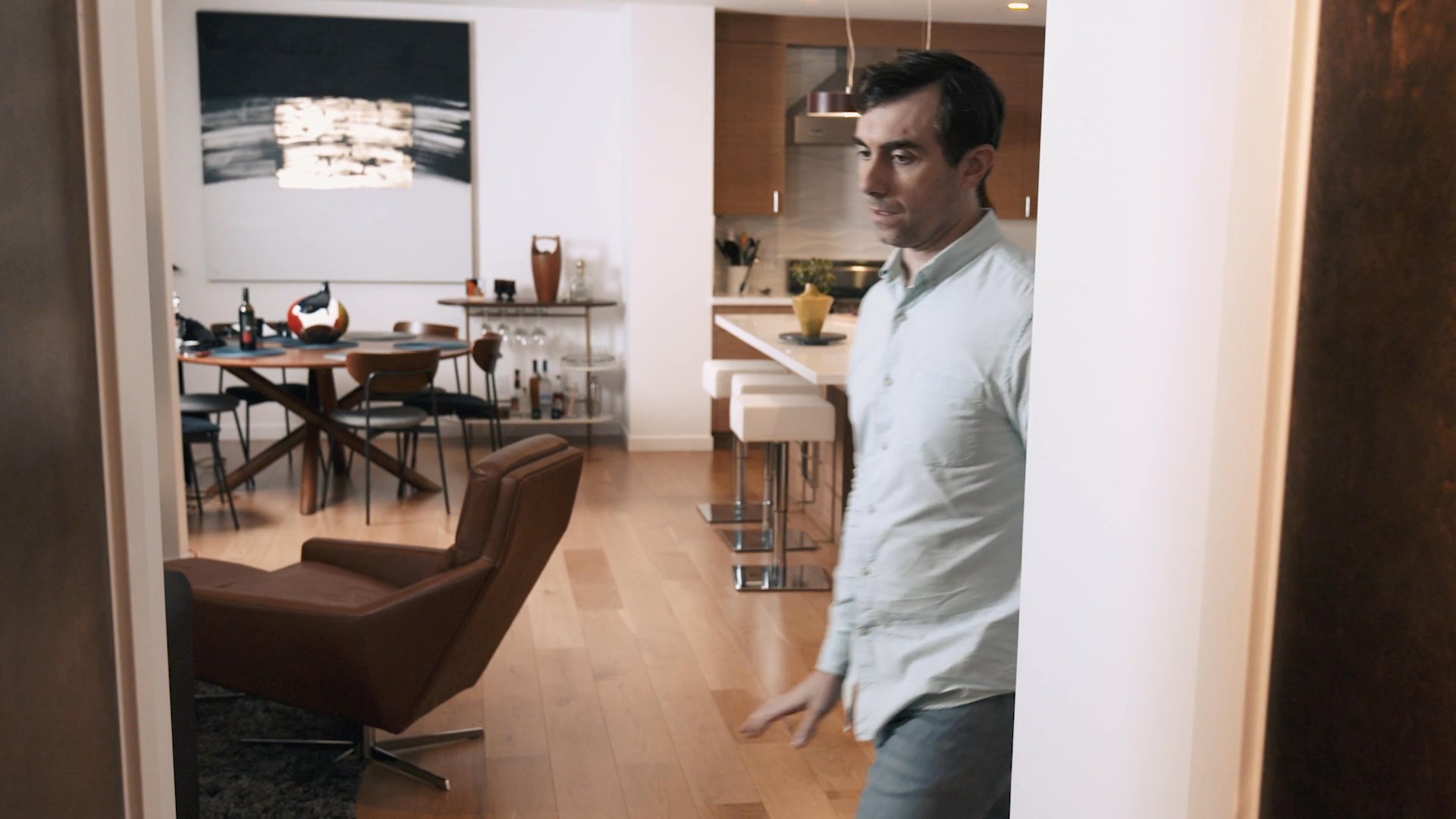 A man arrives at a home and walks through the living area toward a woman standing in the kitchen. She pauses her work to acknowledge his presence.