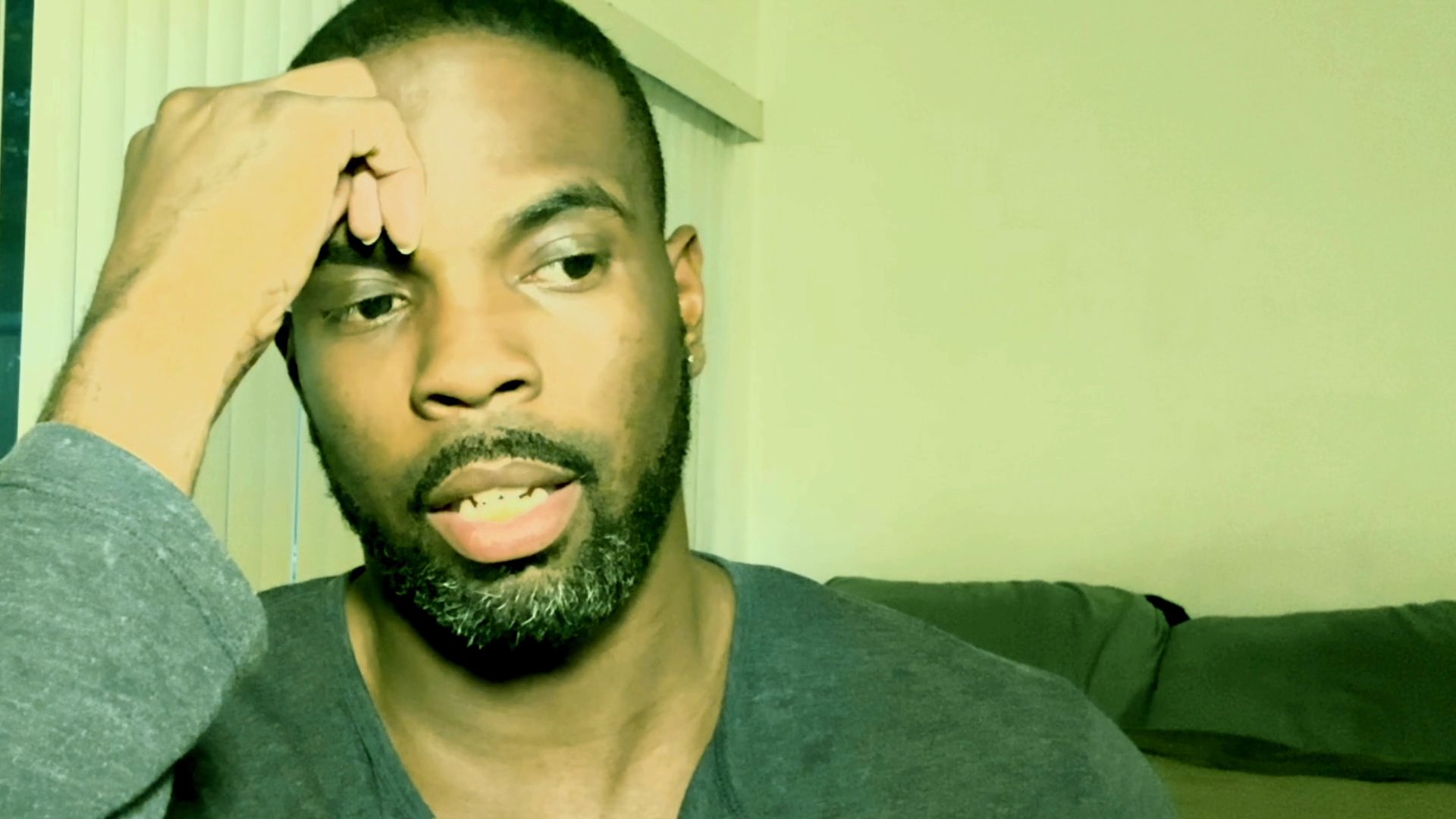 A man speaks directly to the camera while maintaining a relaxed posture with his hand resting near his forehead in an indoor setting.