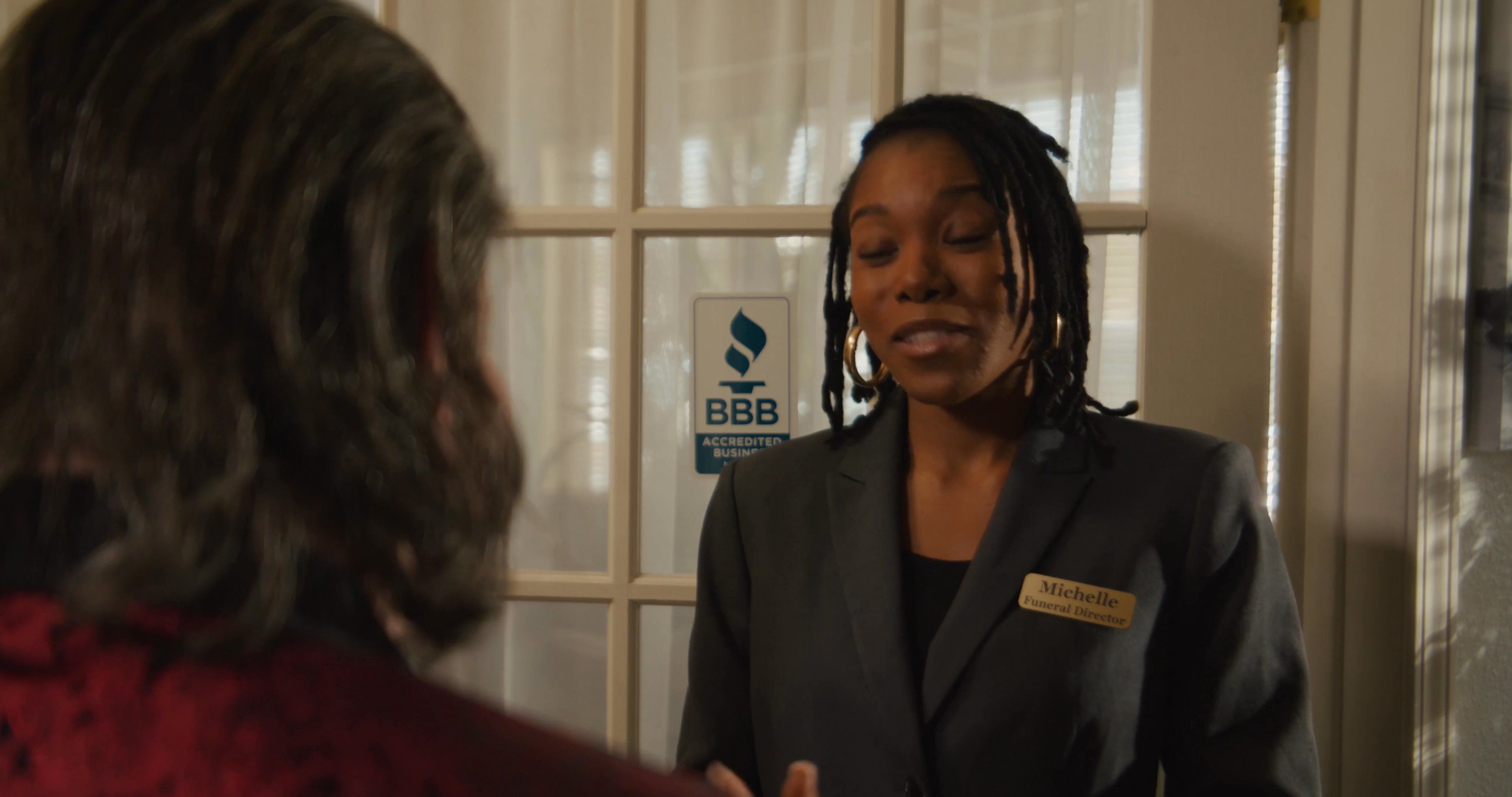 A man in a red formal blazer speaks with a female receptionist in an office setting. They engage in a professional conversation, exchanging pleasantries.