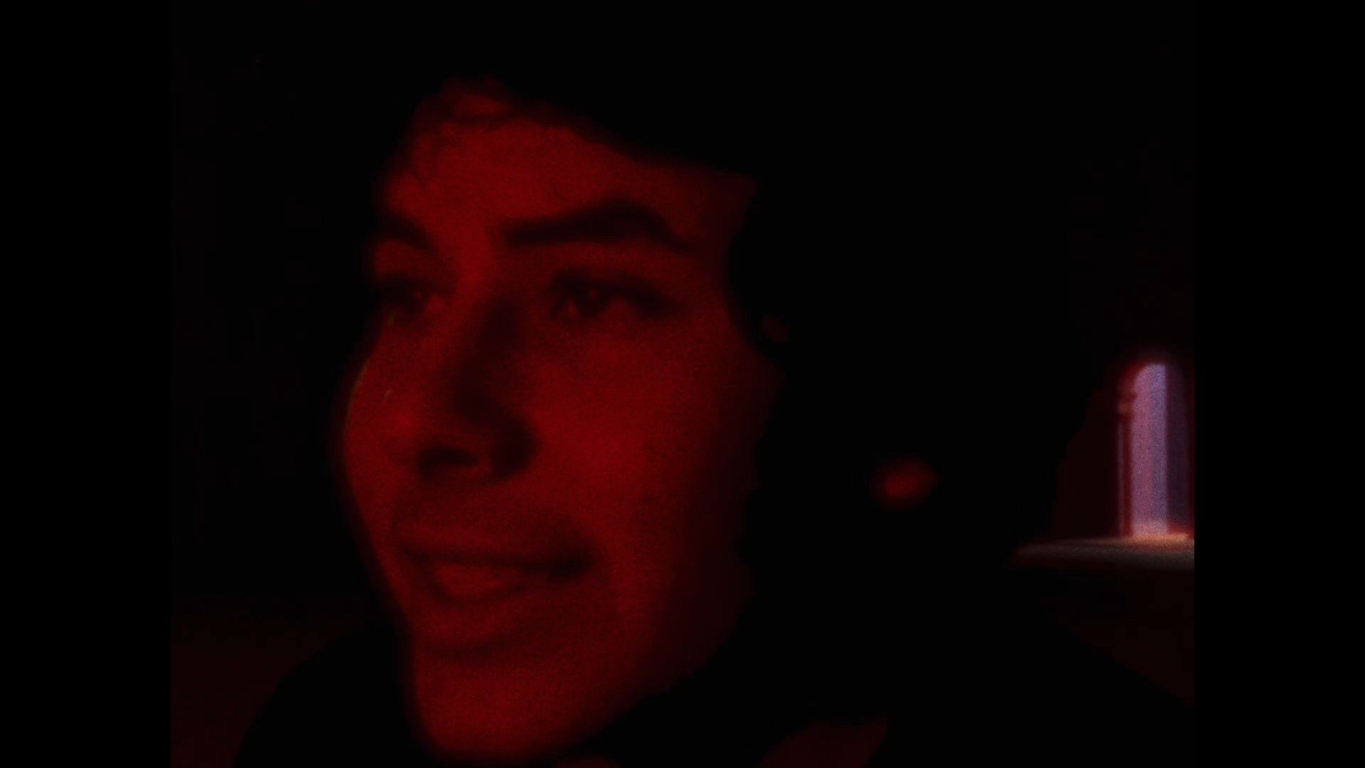 A young man appears in a low-light environment illuminated by red hues, shifting his gaze and facial expressions while speaking. The setting creates a dramatic and intimate atmosphere as he interacts with an unseen person off-camera.