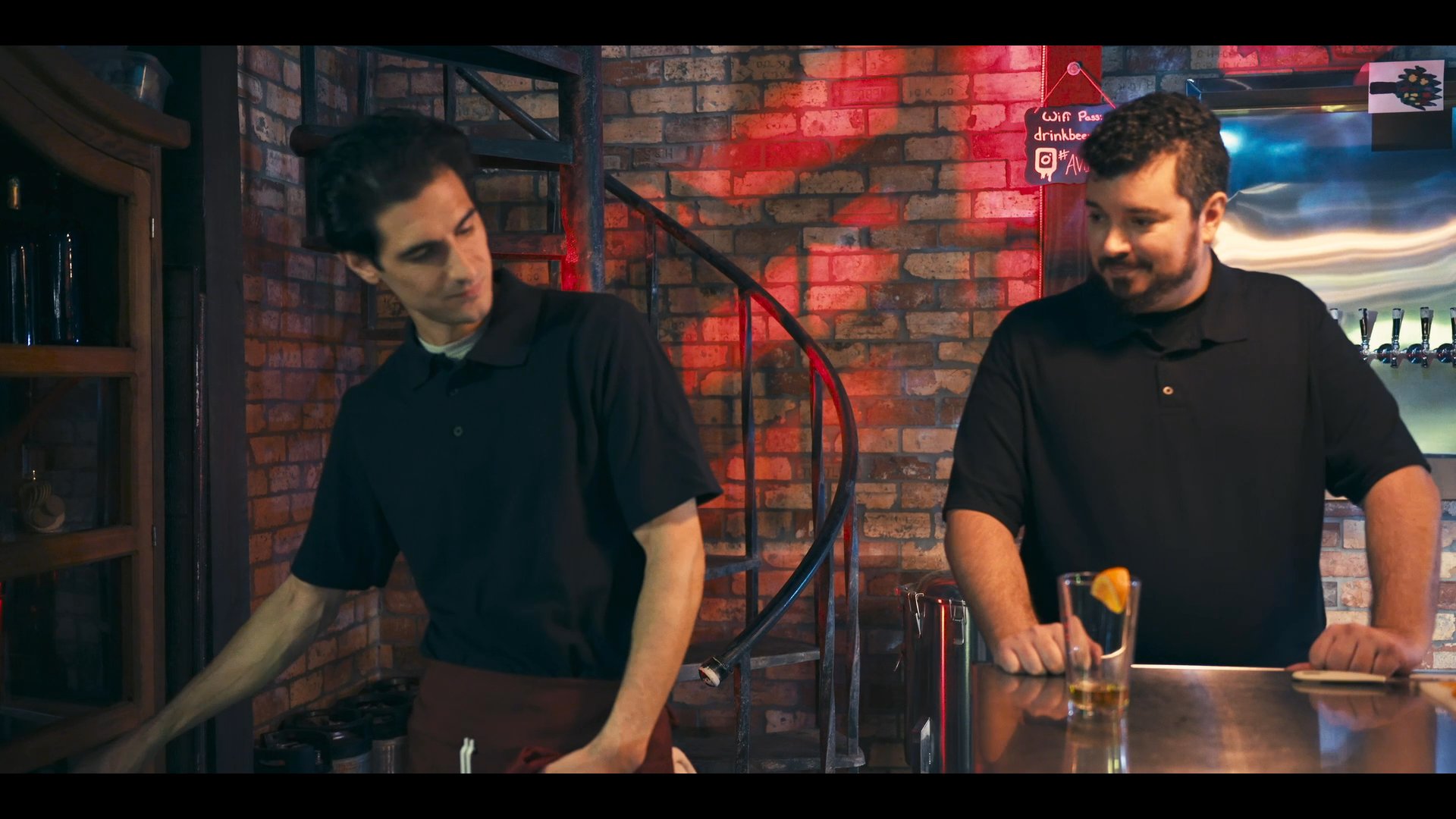 Two bartenders coordinate the preparation and presentation of a drink in a professional bar environment. The primary subject demonstrates cleaning and service techniques with a red napkin while the other provides instructional oversight.