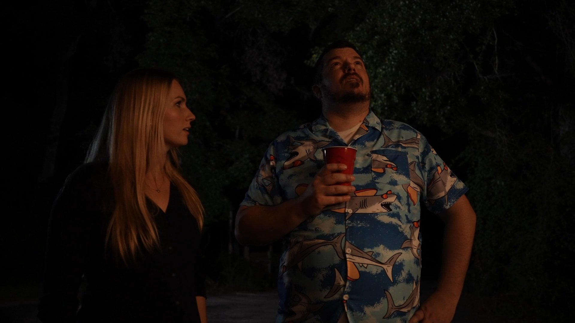 A man and a woman stand together outdoors at night, looking upwards to observe the sky. The man holds a red cup while they both appear engaged in a quiet, evening activity.