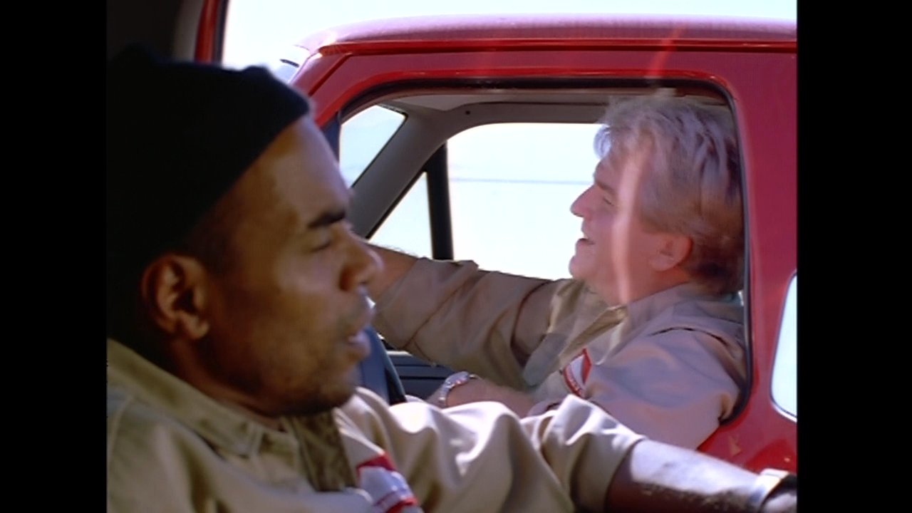 Two men engage in a conversation while sitting inside the cabin of a parked truck. A portable radio is adjusted in the passenger seat as the dialogue continues.