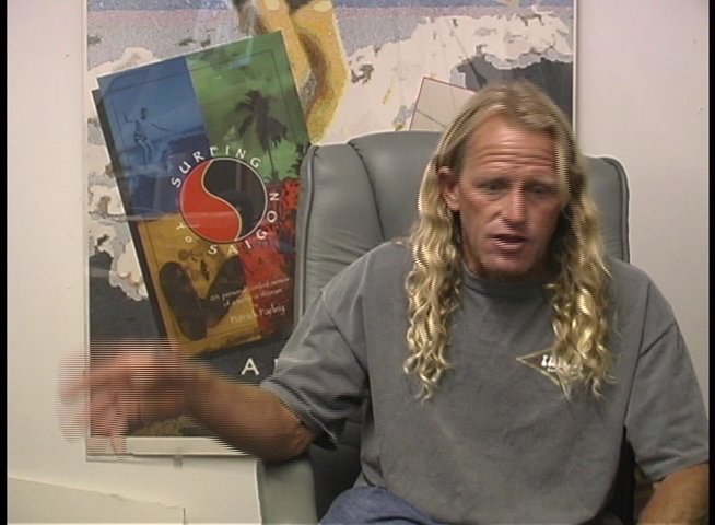 A man with long blond hair sits in an office setting and engages in an animated discussion, using hand gestures to emphasize his points.