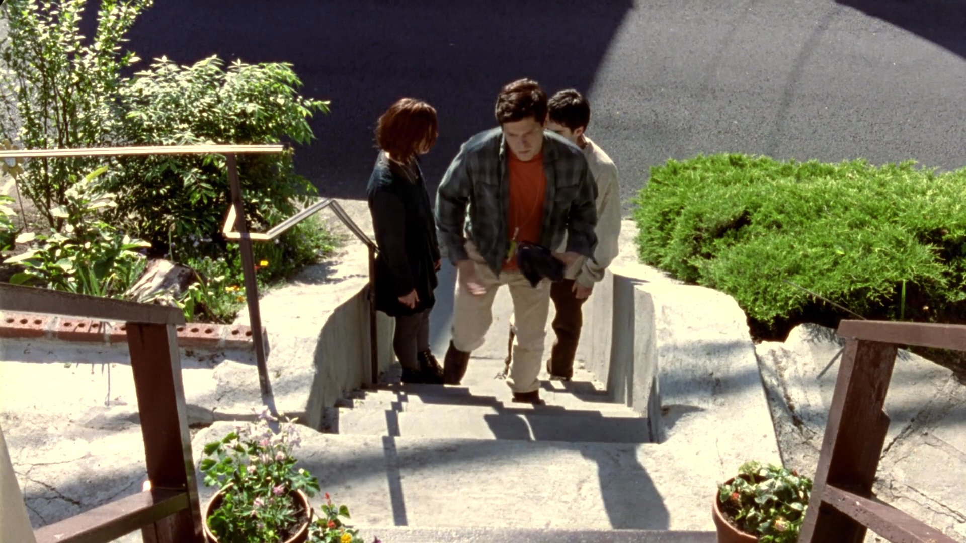 A man and woman converse on residential steps before the man departs, leaving a gift and flowers addressed to a family.