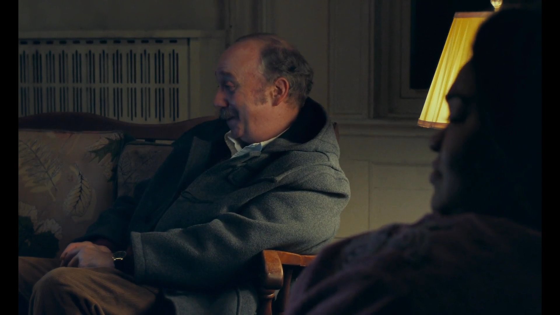 Two individuals engage in a quiet, evening conversation within a dimly lit living room while a television plays in the background.