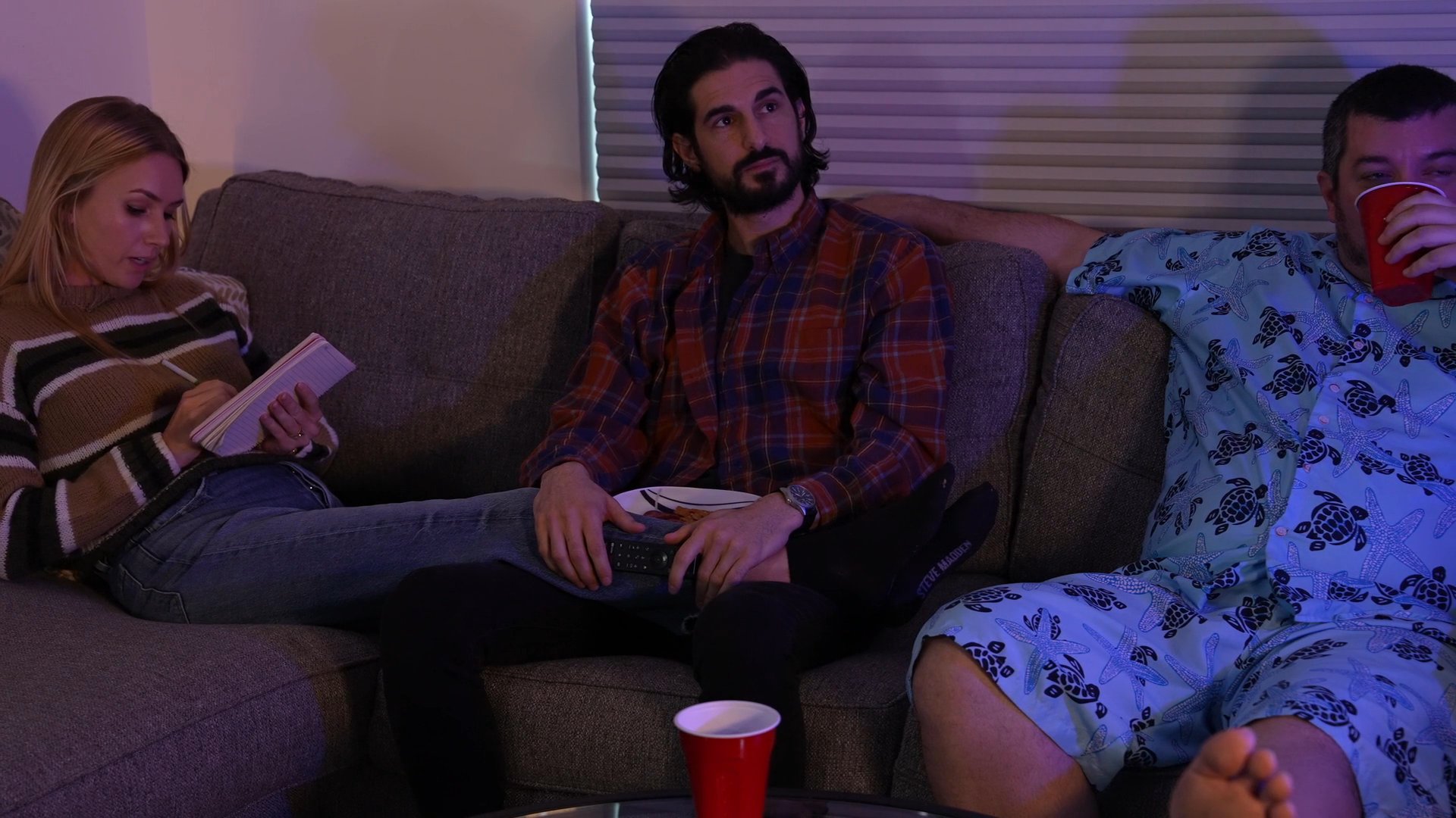 Three individuals engage in a casual discussion while relaxing together on a sectional sofa in a dimly lit living room.
