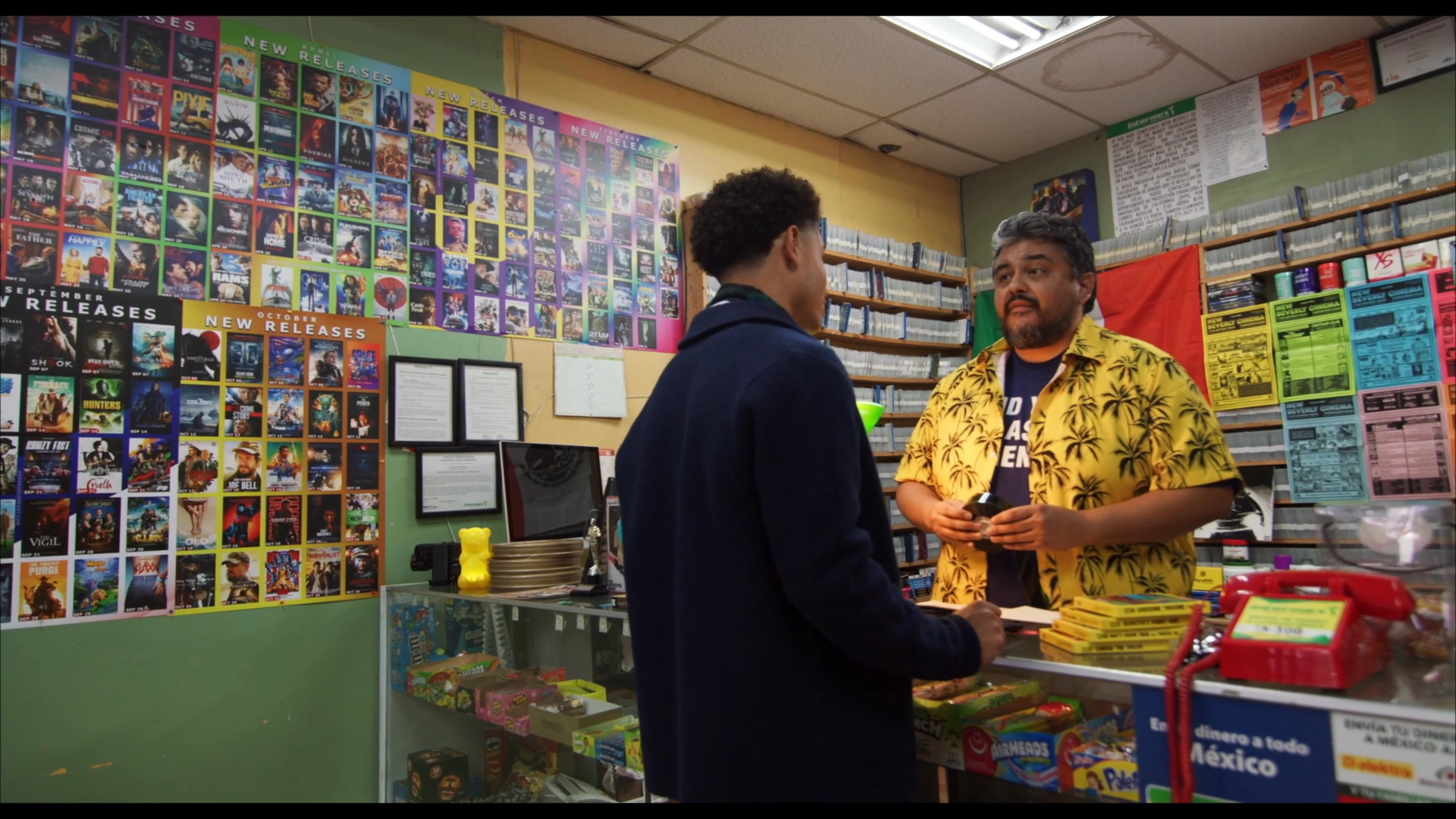 Two young men browse a classic video rental store before one engages in a conversation with the shopkeeper at the counter.