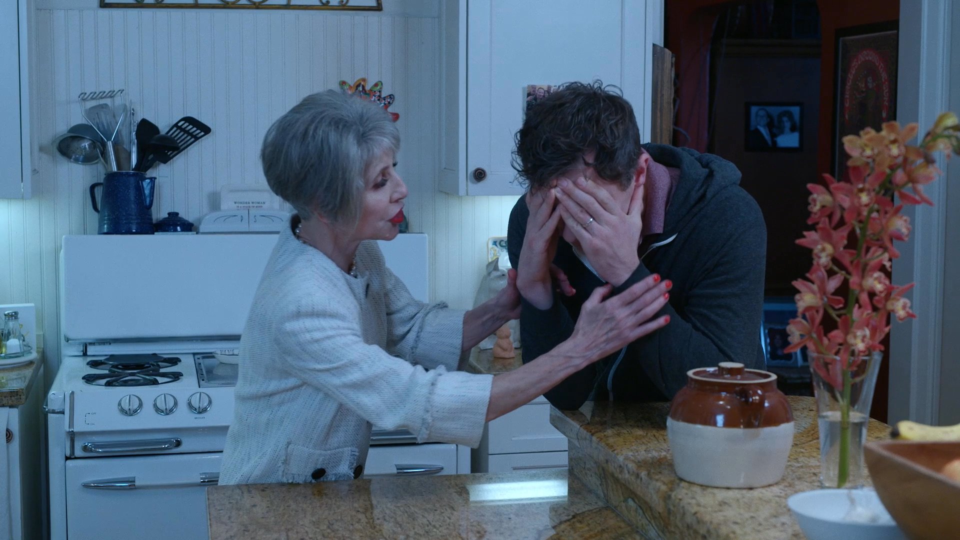 An older woman offers comfort and emotional support to a distressed man who is covering his face in a kitchen setting. She reaches out, takes his hands, and maintains eye contact to provide reassurance.