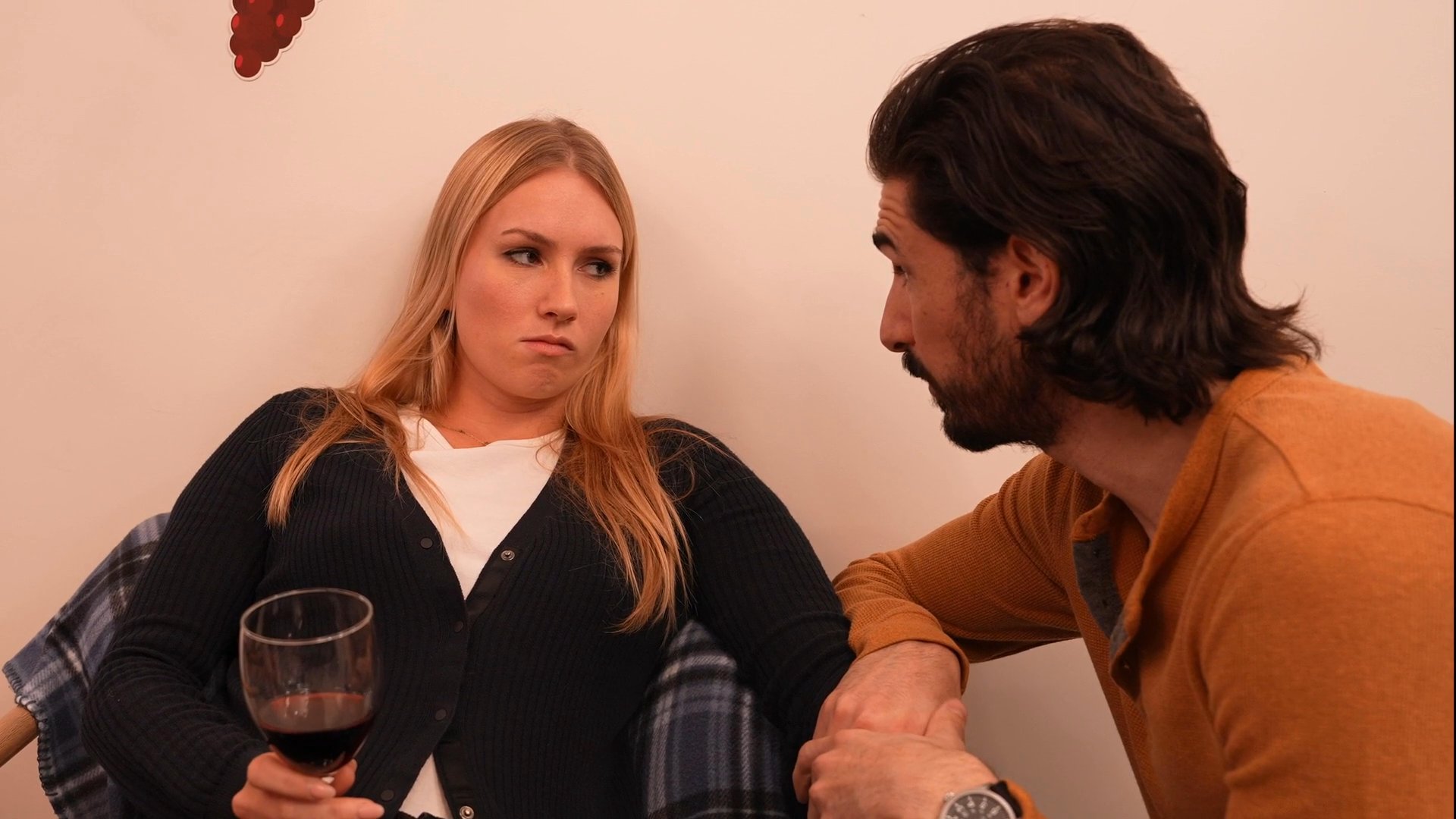 A man engages in a serious conversation with a woman holding a wine glass within a residential living room setting. The scene highlights interpersonal communication dynamics between the two subjects.