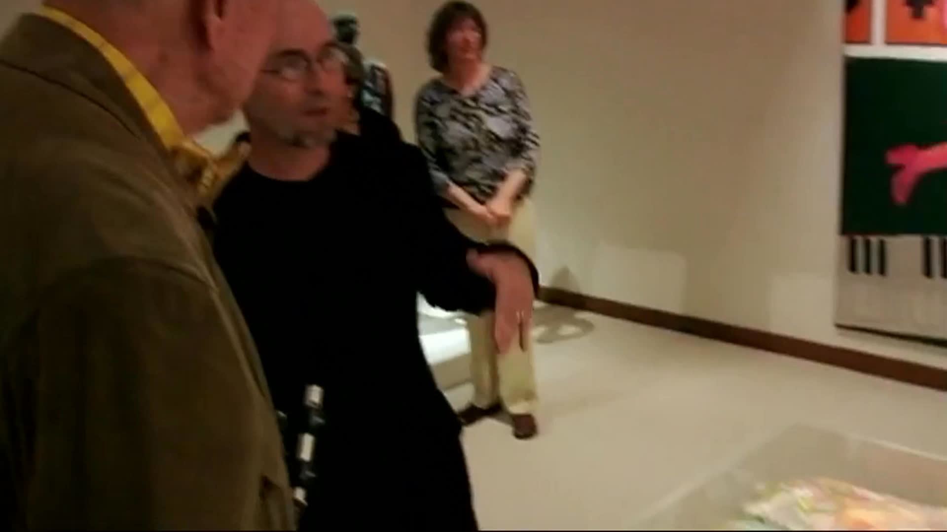 An older man in a green cap and a bald visitor discuss works in a gallery, pausing to examine a vivid, densely layered piece under glass. They continue through the exhibition among colorful paintings and other visitors.