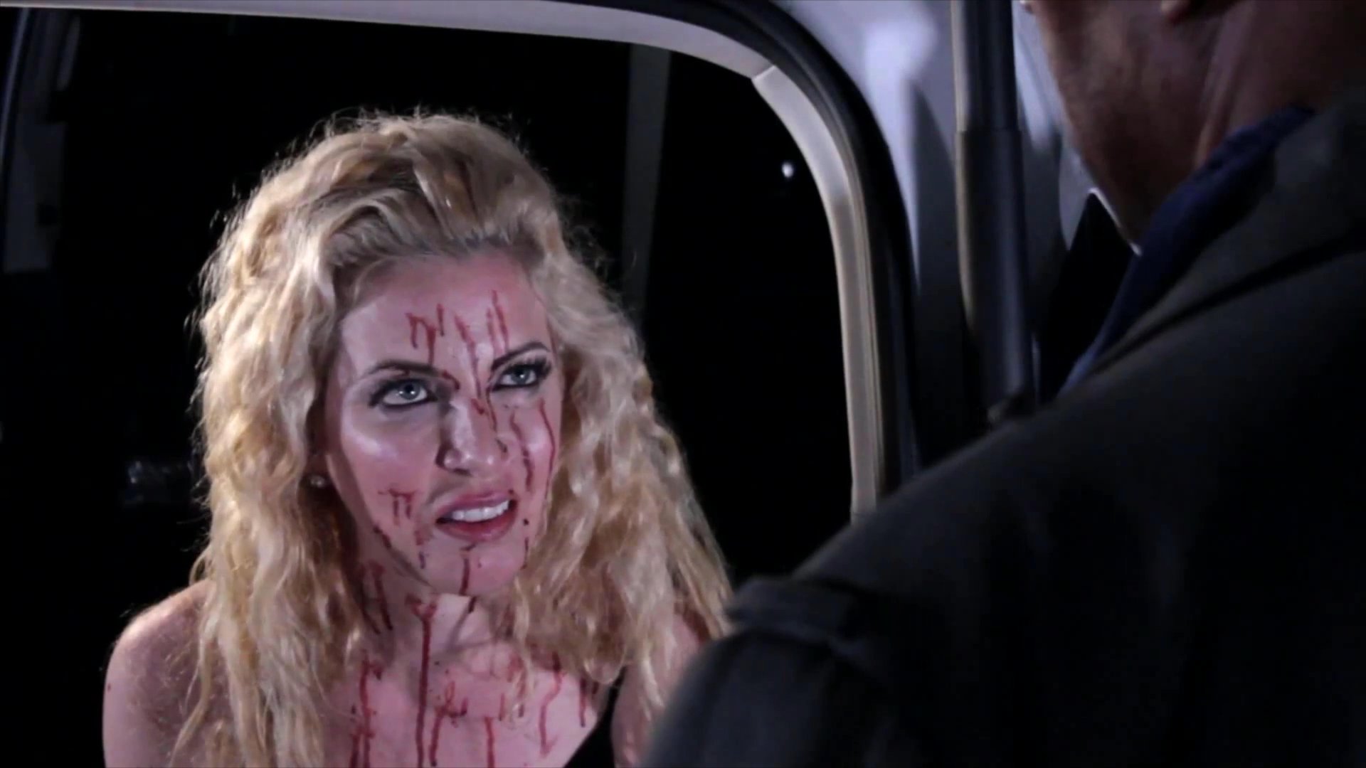 An investigator questions a bloodied woman during an intense, high-stakes confrontation in a dimly lit setting. Their dialogue reveals significant underlying tension as the woman remains distressed.