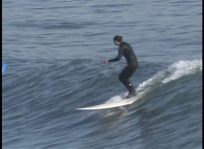 A woman provides commentary before transitioning to footage of surfers riding waves in the ocean while wearing wetsuits.