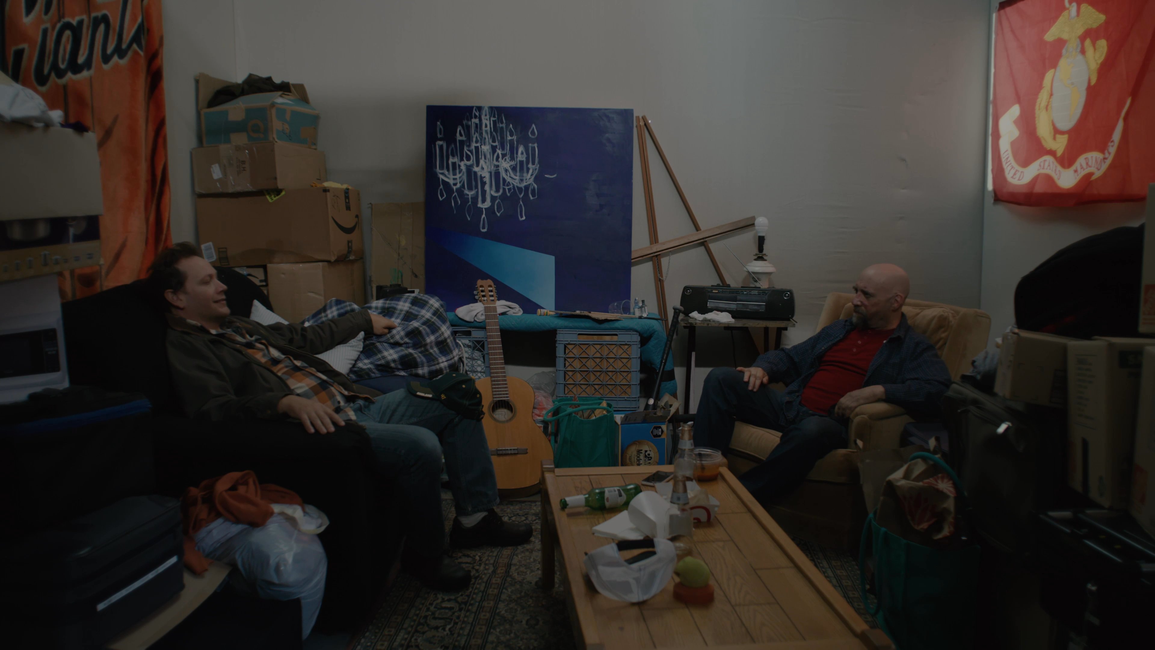 Two men engage in a conversational exchange while seated in a cluttered, informal living room environment. They alternate between speaking and listening as they sit across from one another.
