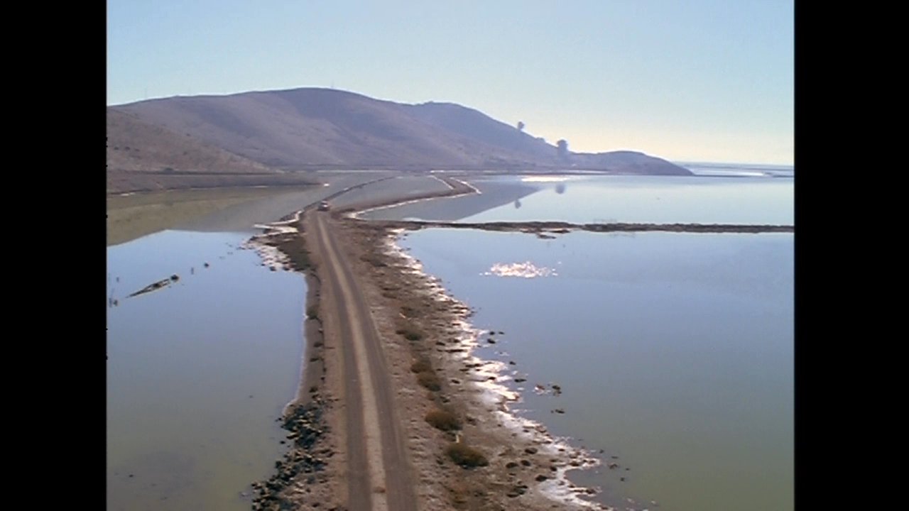 A field technician performs maintenance and gathers environmental data near a remote salt flat infrastructure site. The scene transitions from close-up technical work to a wide aerial view of the isolated landscape.