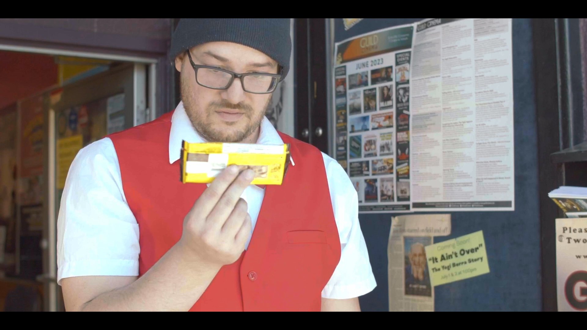 A theatre employee in a red vest picks up a dropped candy bar from the sidewalk and examines it before heading into the cinema. Inside, two patrons sit in the theatre seats as the employee observes the surroundings.