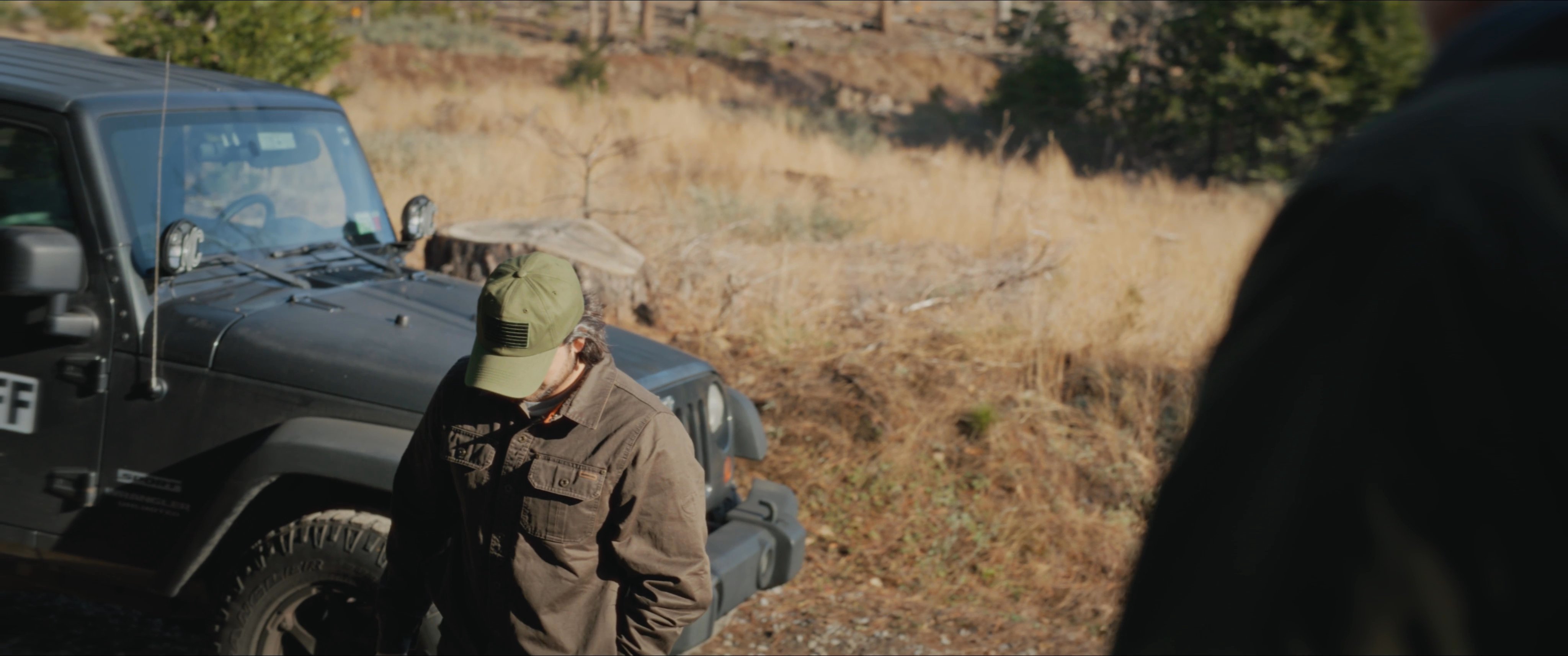 A man in a brown jacket and baseball cap converses with a sheriff standing near a marked patrol vehicle in a forested area.