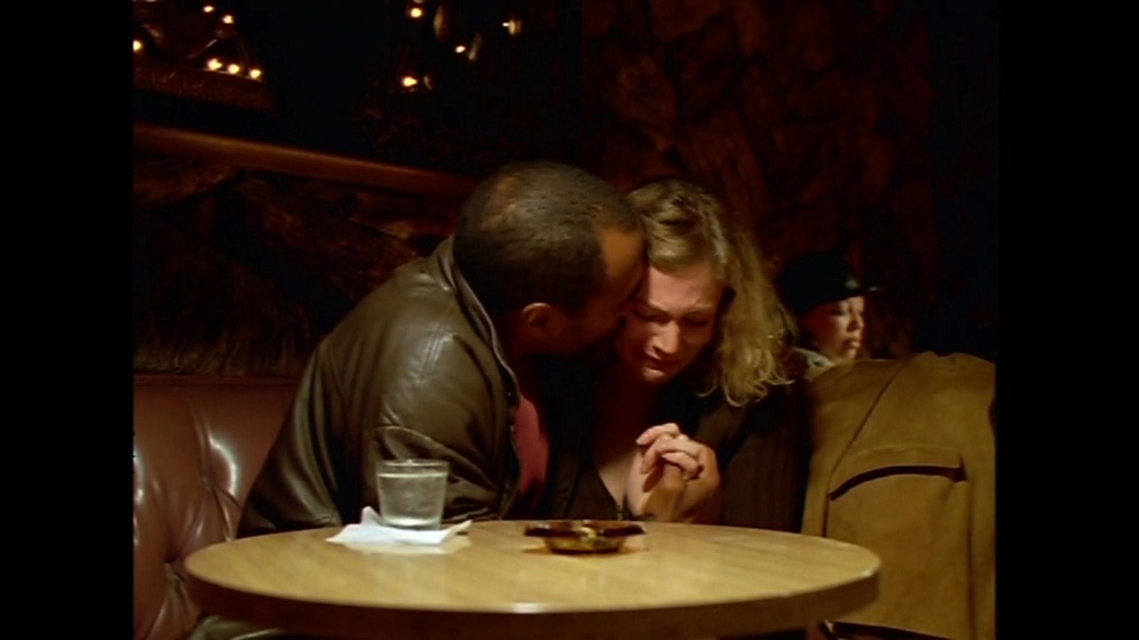 A man and woman share a tender, private conversation while seated together in a dim, dimly lit restaurant booth.