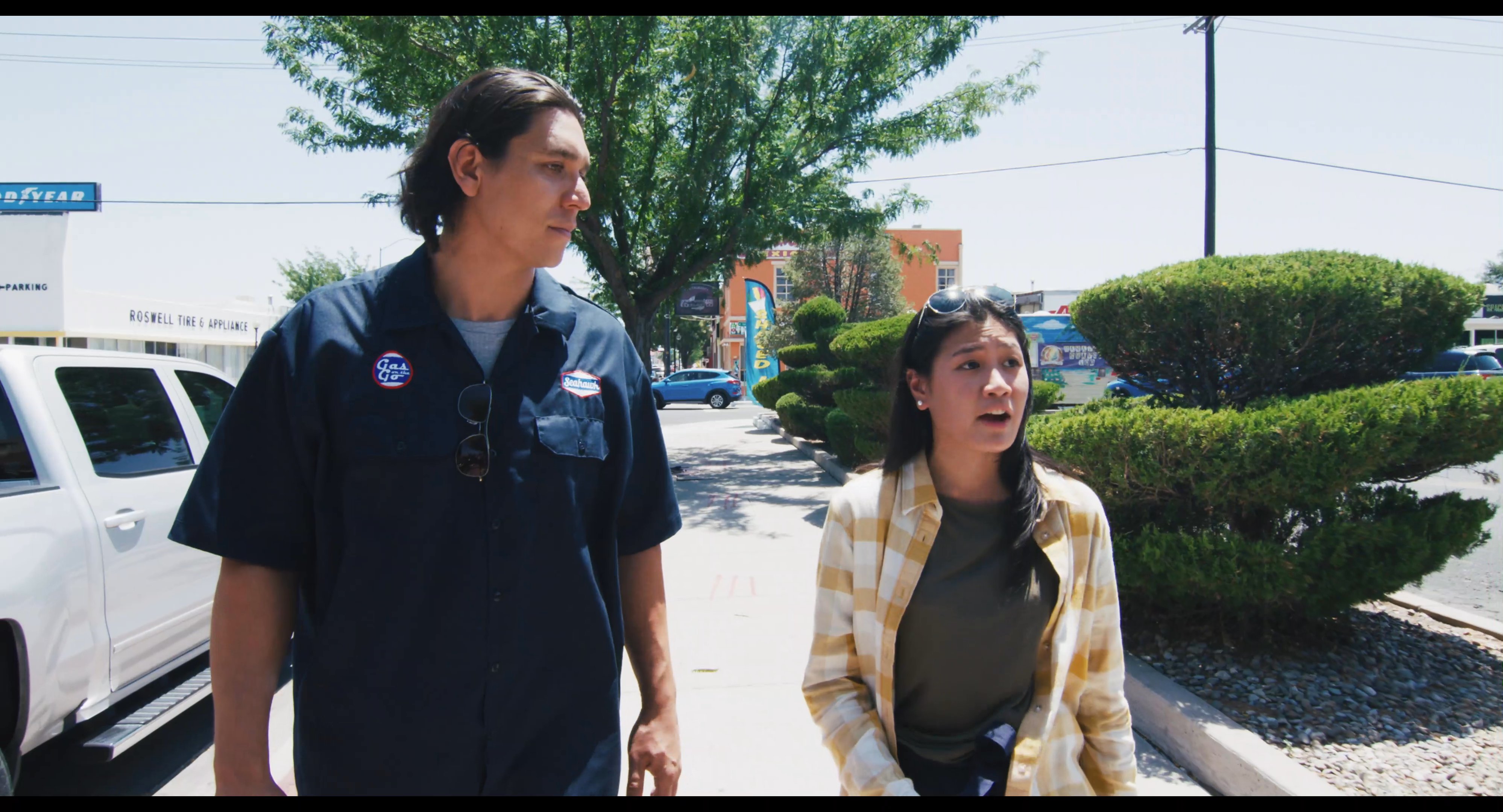 A man and a woman engage in a continuous, active conversation while walking through a sunny parking lot.