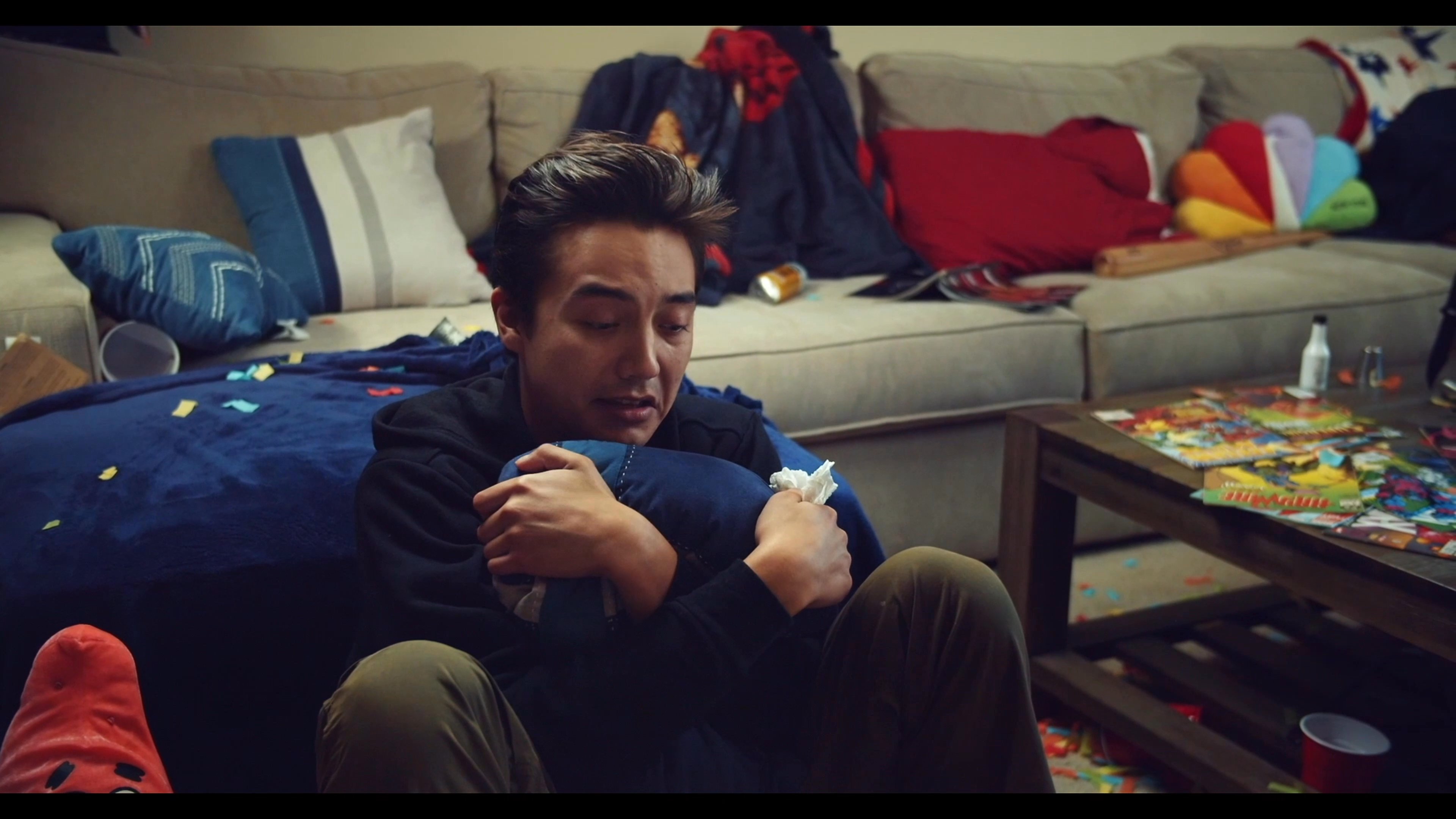 Two young men engage in a tense, intimate conversation within a shared living space, with one person visibly distressed on the floor while the other addresses him.