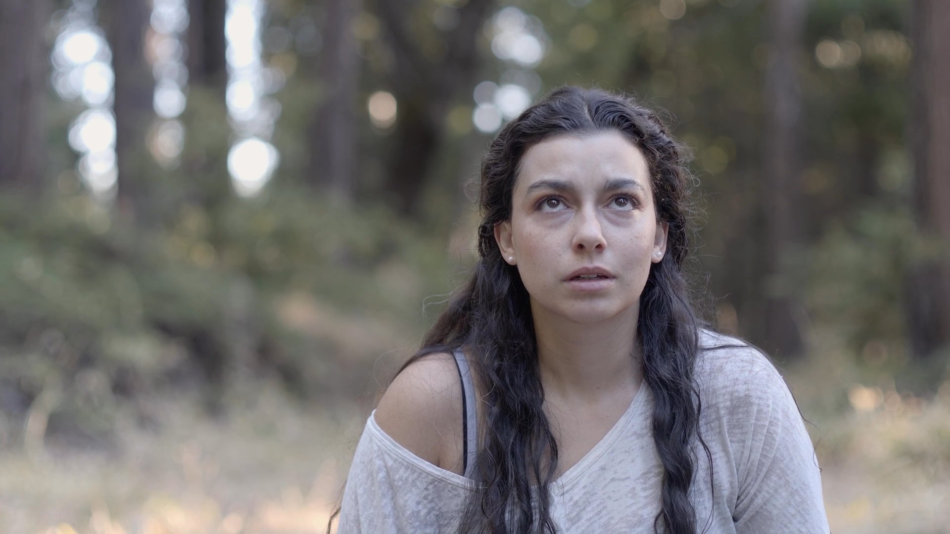 A woman sits in a tranquil forest setting, shifting her gaze slowly from the ground to the towering trees above. The scene captures a moment of quiet introspection amidst natural surroundings.