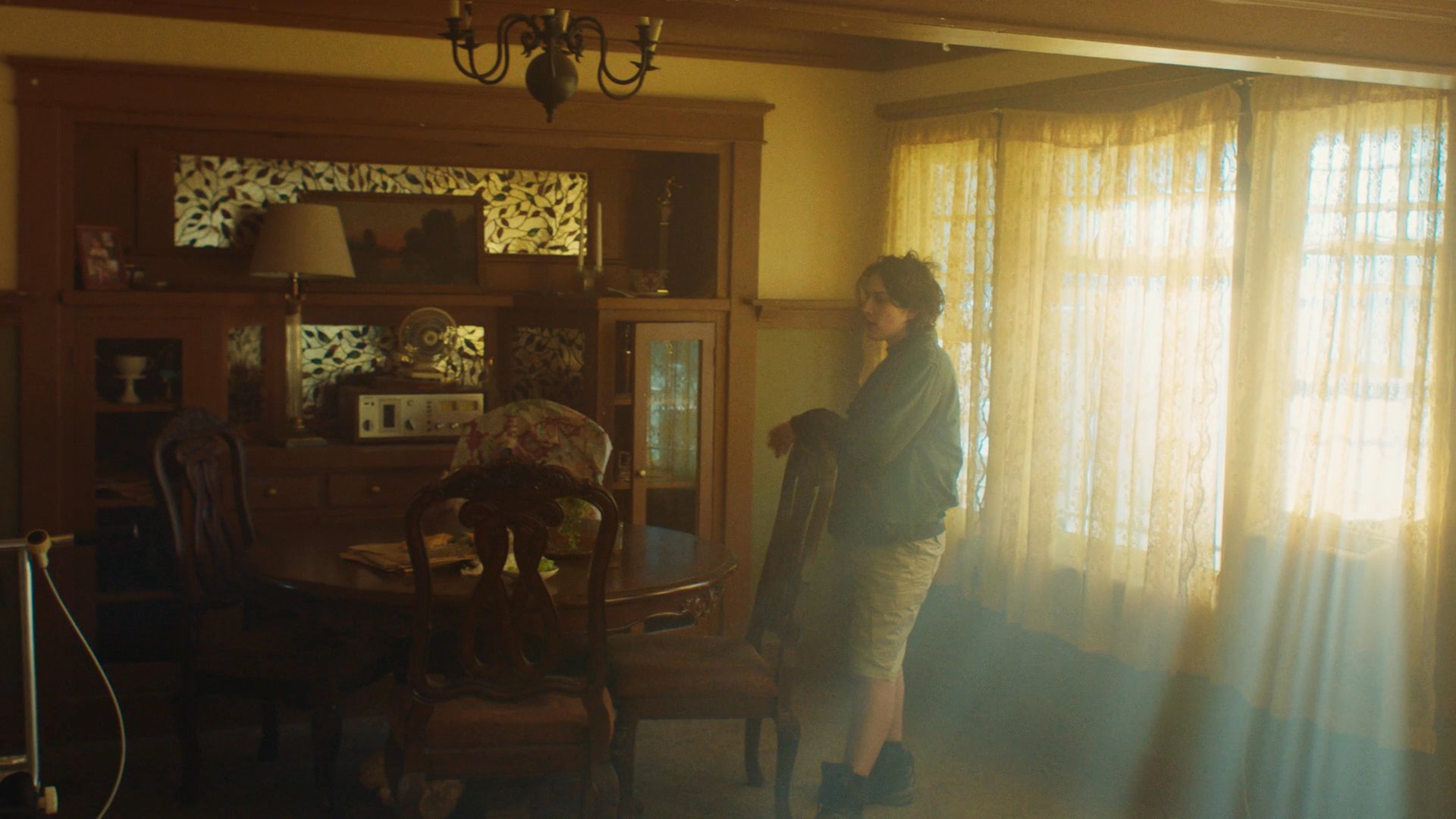 A woman navigates a tense, volatile household environment marked by substance use and underlying interpersonal strain. The scene captures the fractured dynamics between individuals within a sunlit, cluttered domestic space.