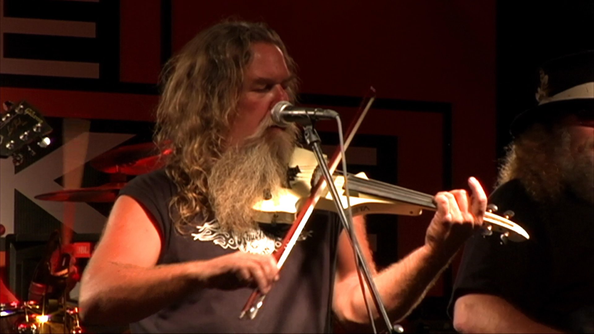 A long-haired fiddler performs on stage with his band before shifting to scenes of bikers riding in a group along a vast, open highway.