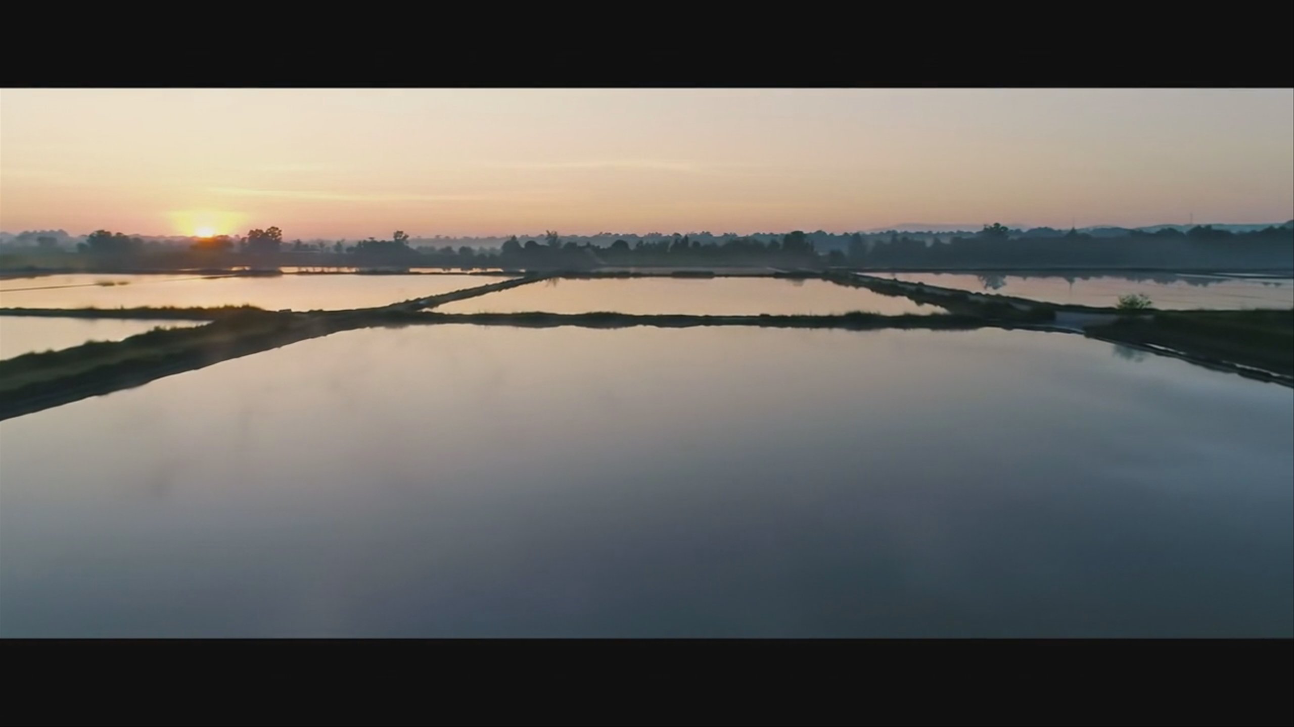 An aerial glide over terraced salt flats mirroring a pastel sunrise sky.