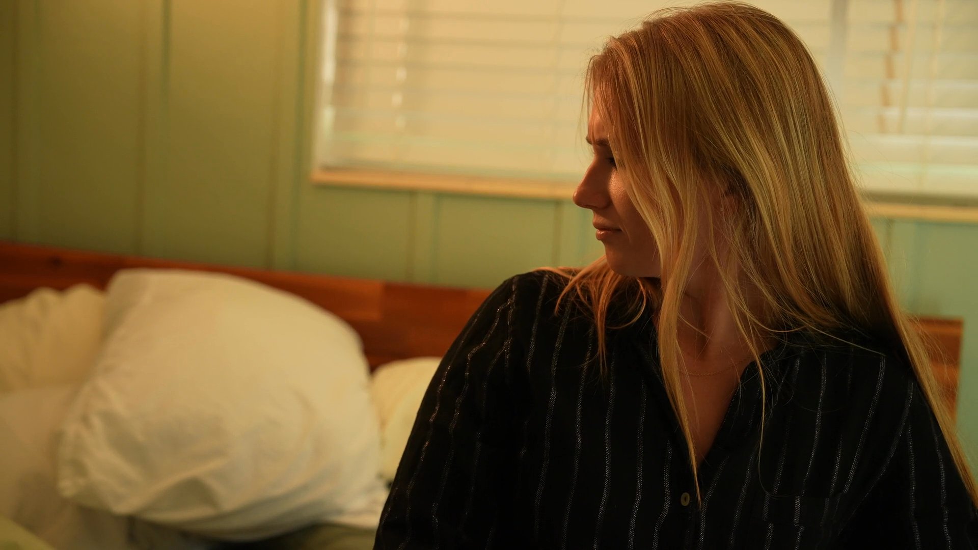 A woman sits on a bed and observes as a man enters the room and gestures towards her, initiating a conversation in a domestic setting.