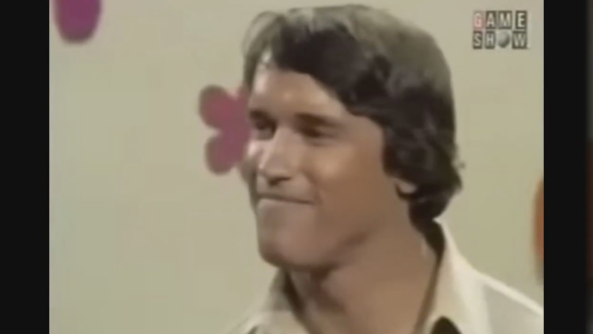 Celebrity guests participate in a televised 1970s game show set against a colorful floral-patterned background. The host moderates the panel as contestants and famous figures engage with the audience.