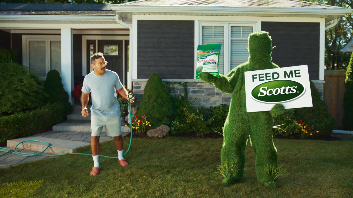 Film still: the lawn character holding Scotts Turf Builder and a feed-me sign beside the homeowner