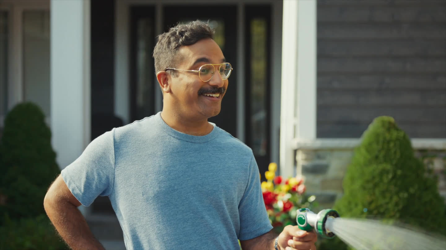 Film still: a smiling homeowner holding a garden hose on a lawn