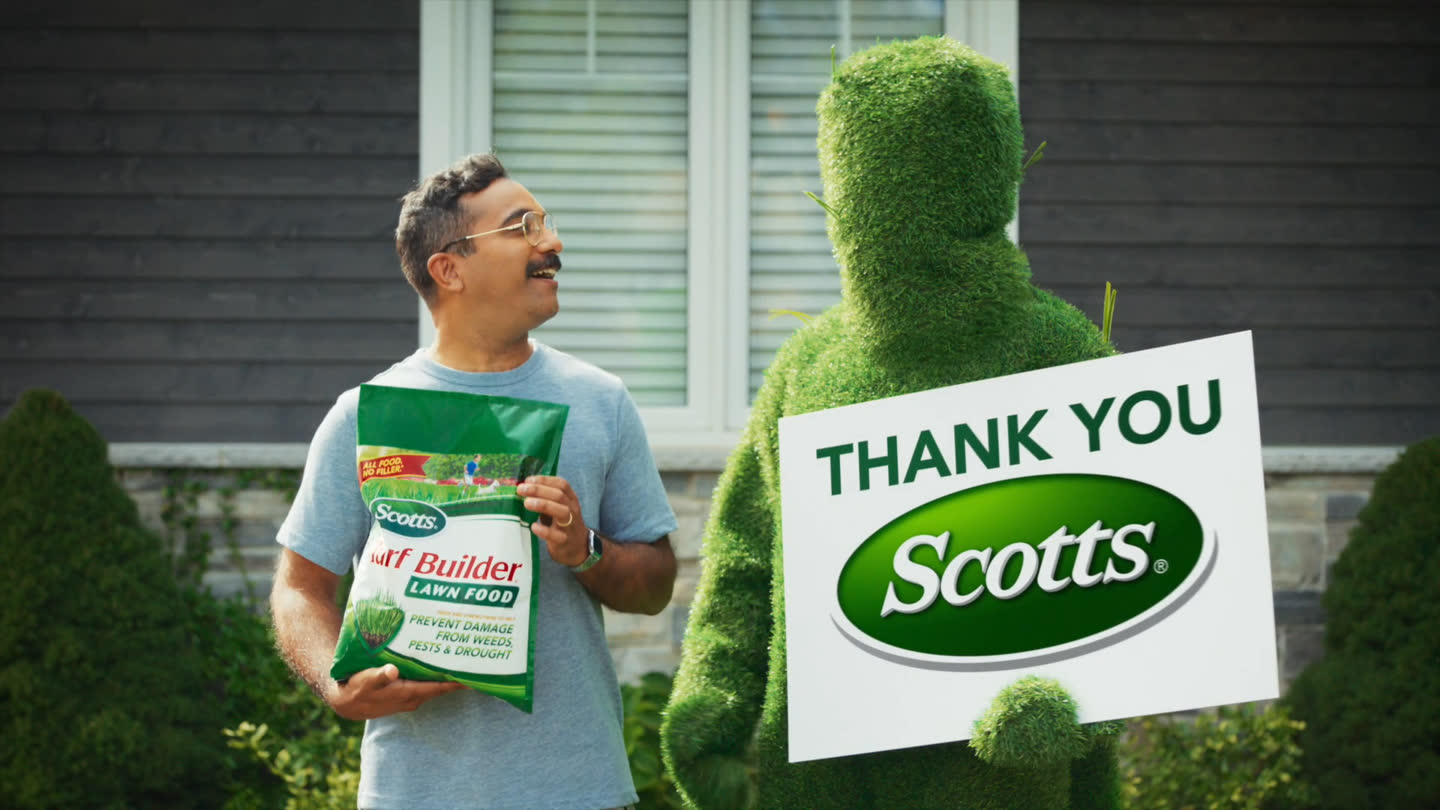 Film still: a smiling homeowner holding Scotts Turf Builder beside the lawn character and its thank-you sign