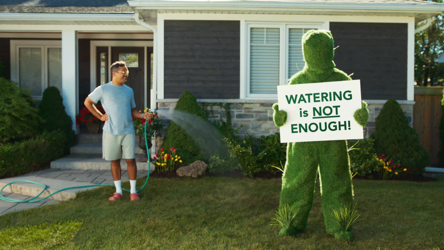 Film still: a homeowner looking at the lawn-costumed character holding a watering sign
