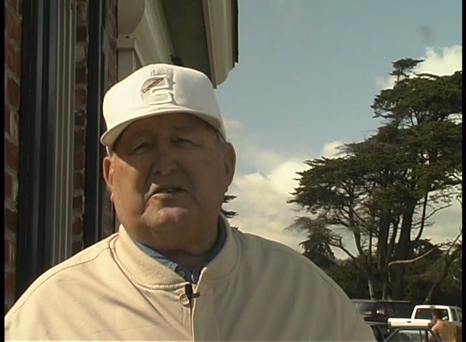An elderly man wearing headphones sits outdoors while sharing personal experiences in an interview setting. The scene transitions to a man in a cap speaking directly to the camera, providing context or narrative reflection.