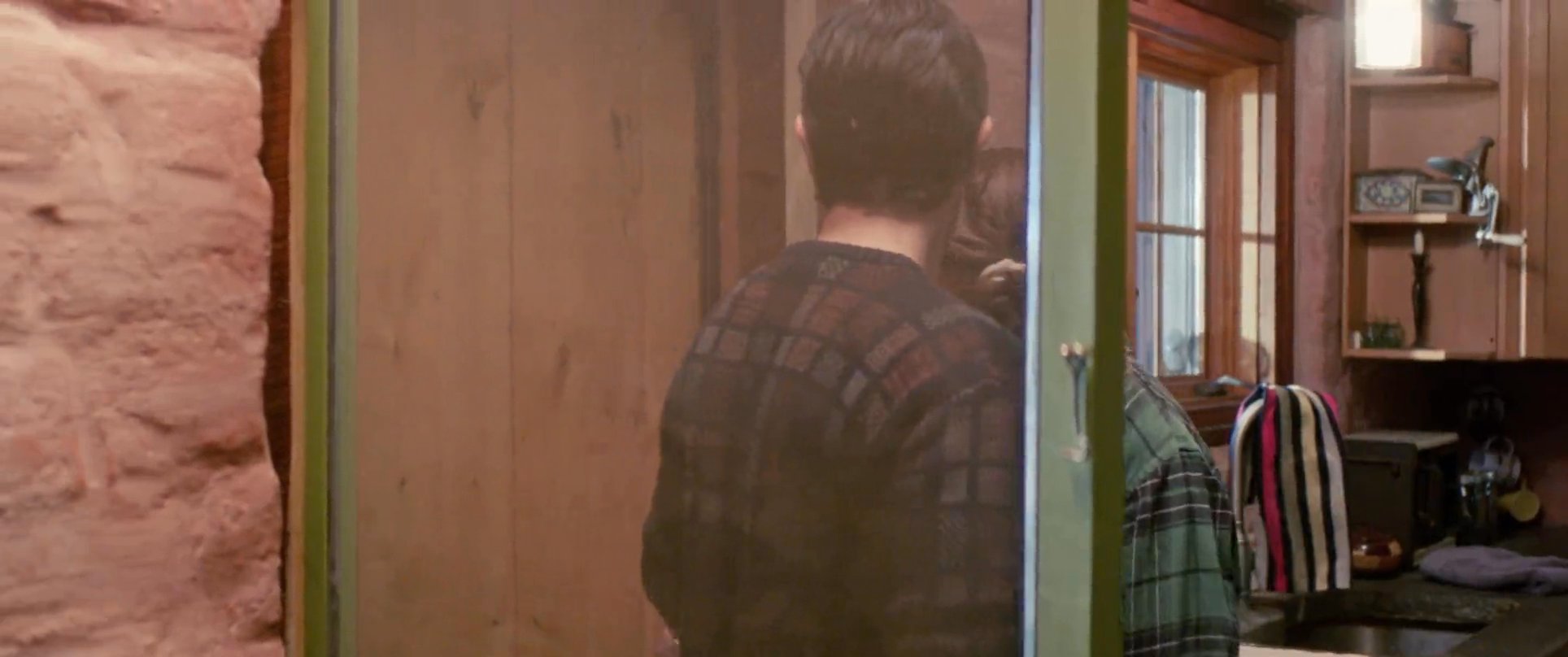 Two men engage in conversation at a doorway before one welcomes a woman into the home with a warm embrace. The scene captures an intimate moment of arrival and interpersonal connection in a residential setting.