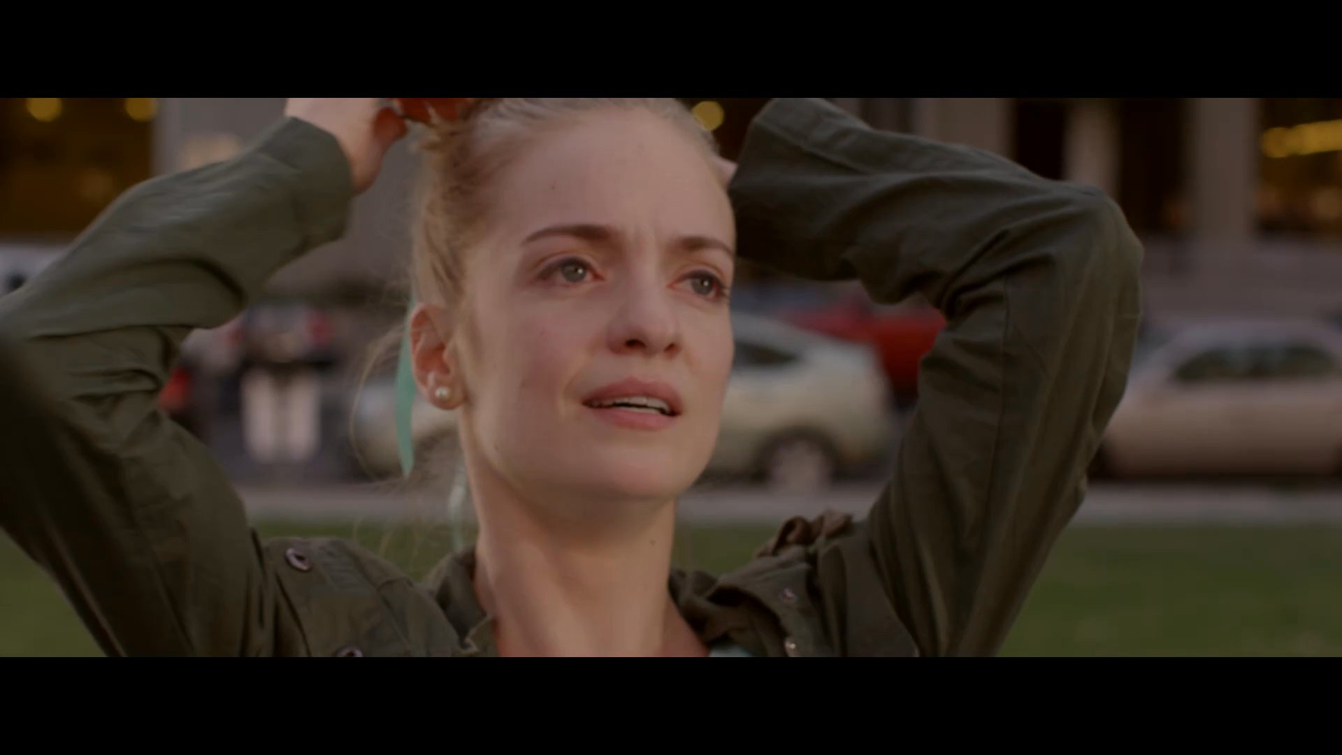 A young woman walks through an urban park before sitting on a bench, overcome by intense emotional distress as she pulls at her hair.
