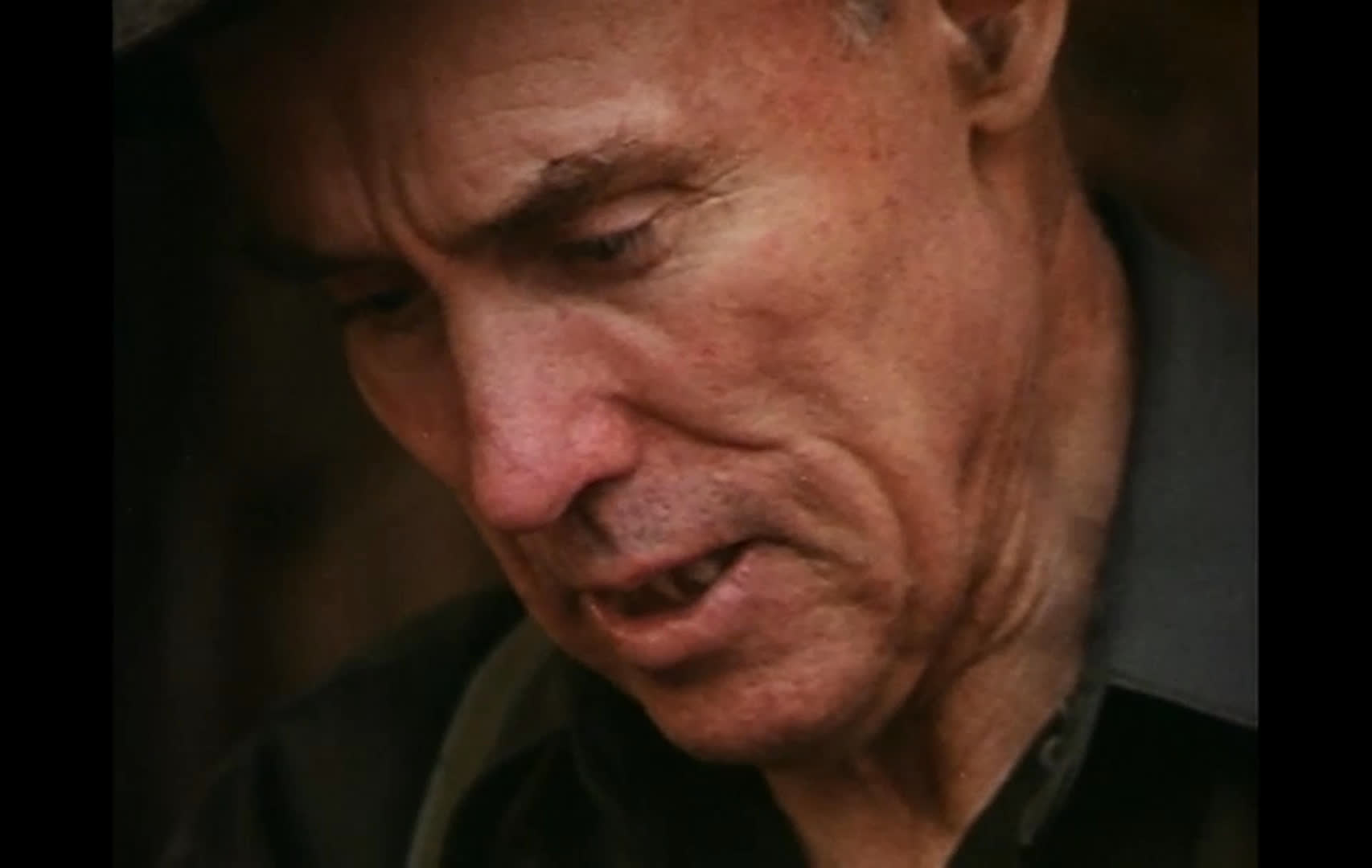 A man in a cowboy hat talks about early-morning life on the range and making coffee over a fire. In a rustic kitchen, he leans over a breakfast table while a woman drinks from a cup.