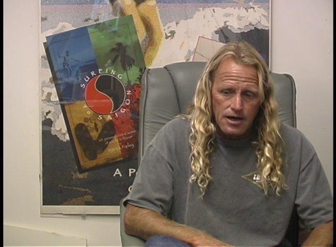 A long-haired man sits in a professional office environment, discussing surfing and surf culture while gesturing to emphasize his points.