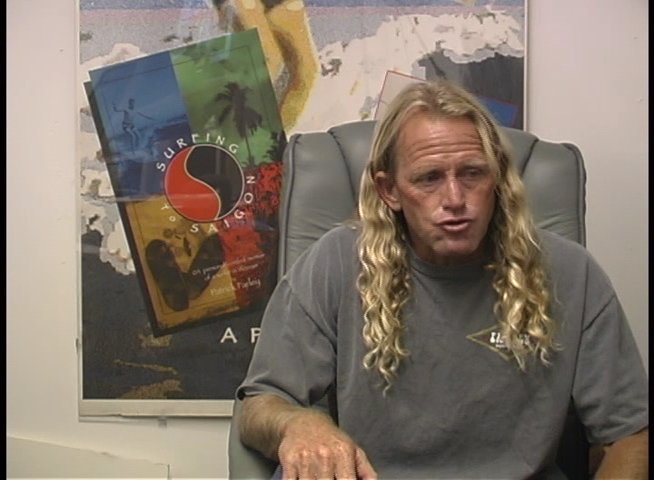 A man with long blonde hair discusses his experiences while sitting in a studio chair in front of a framed poster. He uses expressive hand gestures to emphasize his points during the interview.