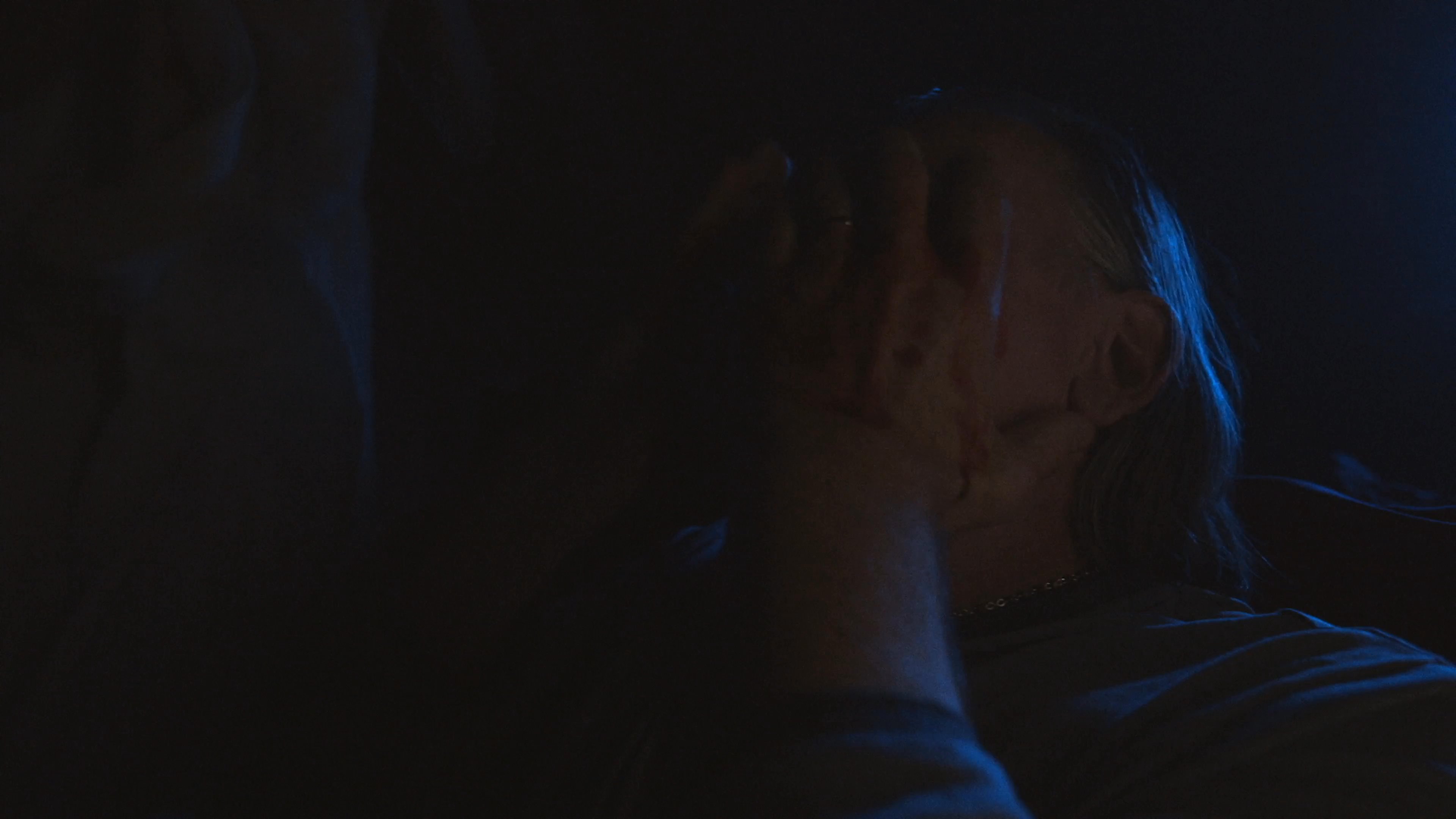 A distressed woman encounters a man suffering from a facial injury in a dimly lit attic. She attempts to provide care by applying a bandage to his bleeding face amidst a tense and dramatic atmosphere.