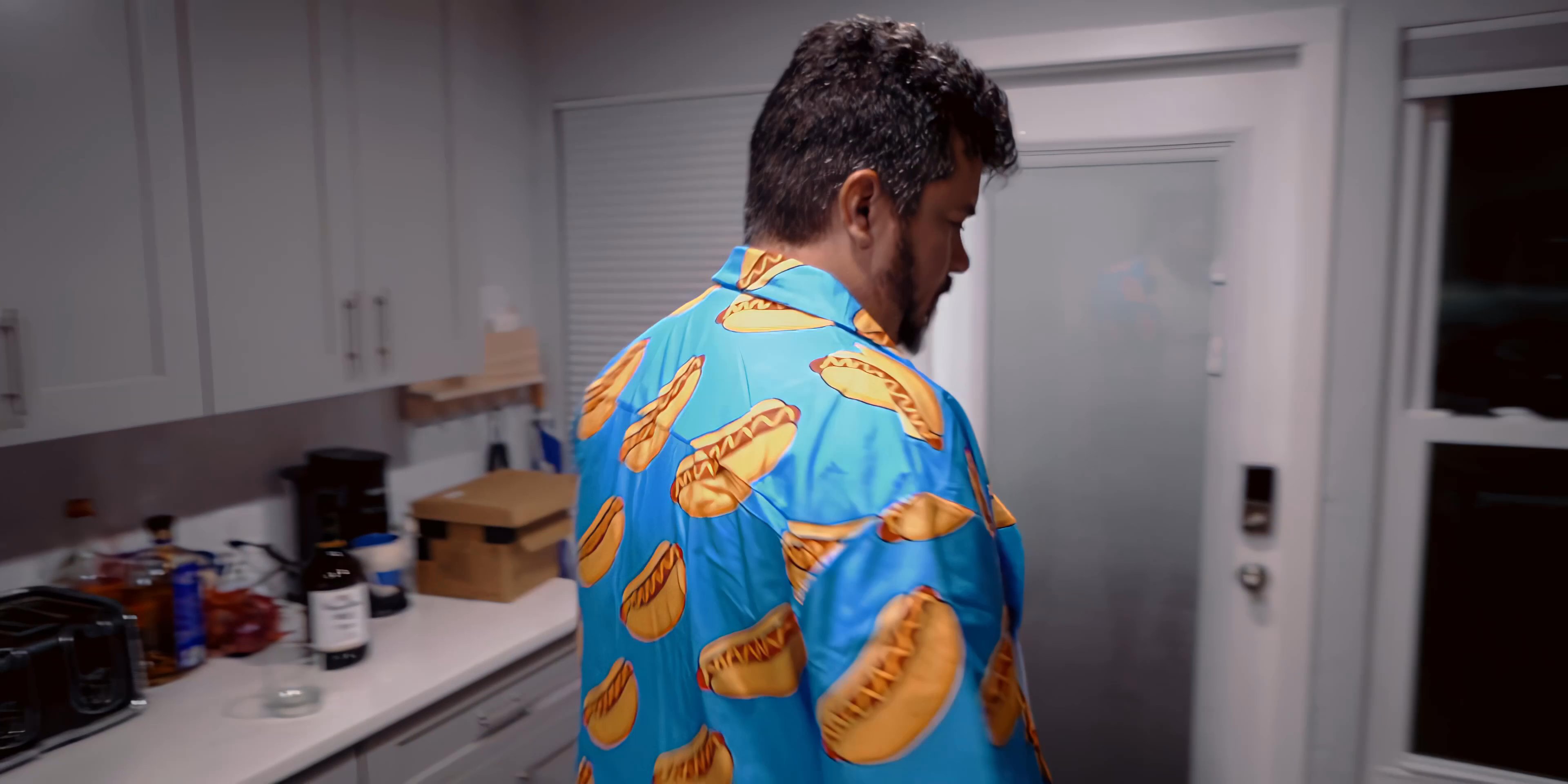 A man wearing a patterned shirt opens his refrigerator to look for food before walking into a dimly lit living area. He observes the room and approaches a recliner in the home.