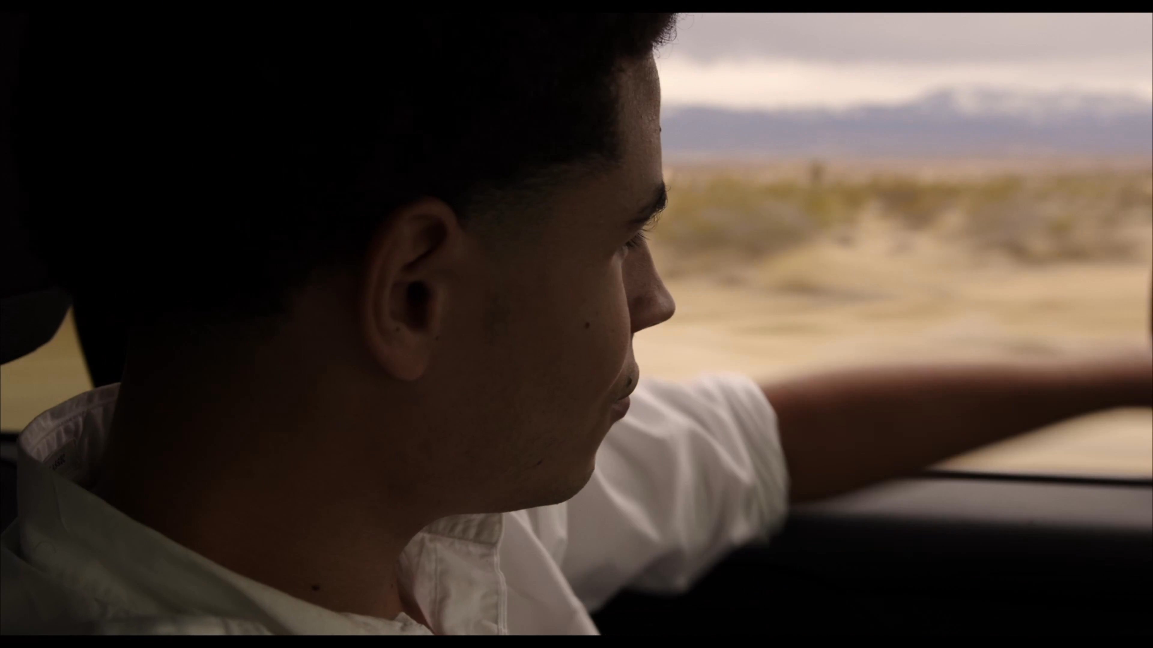 Two friends prepare their vehicle in a parking lot before embarking on a desert journey, where one passenger wears a blindfold while looking out the car window.