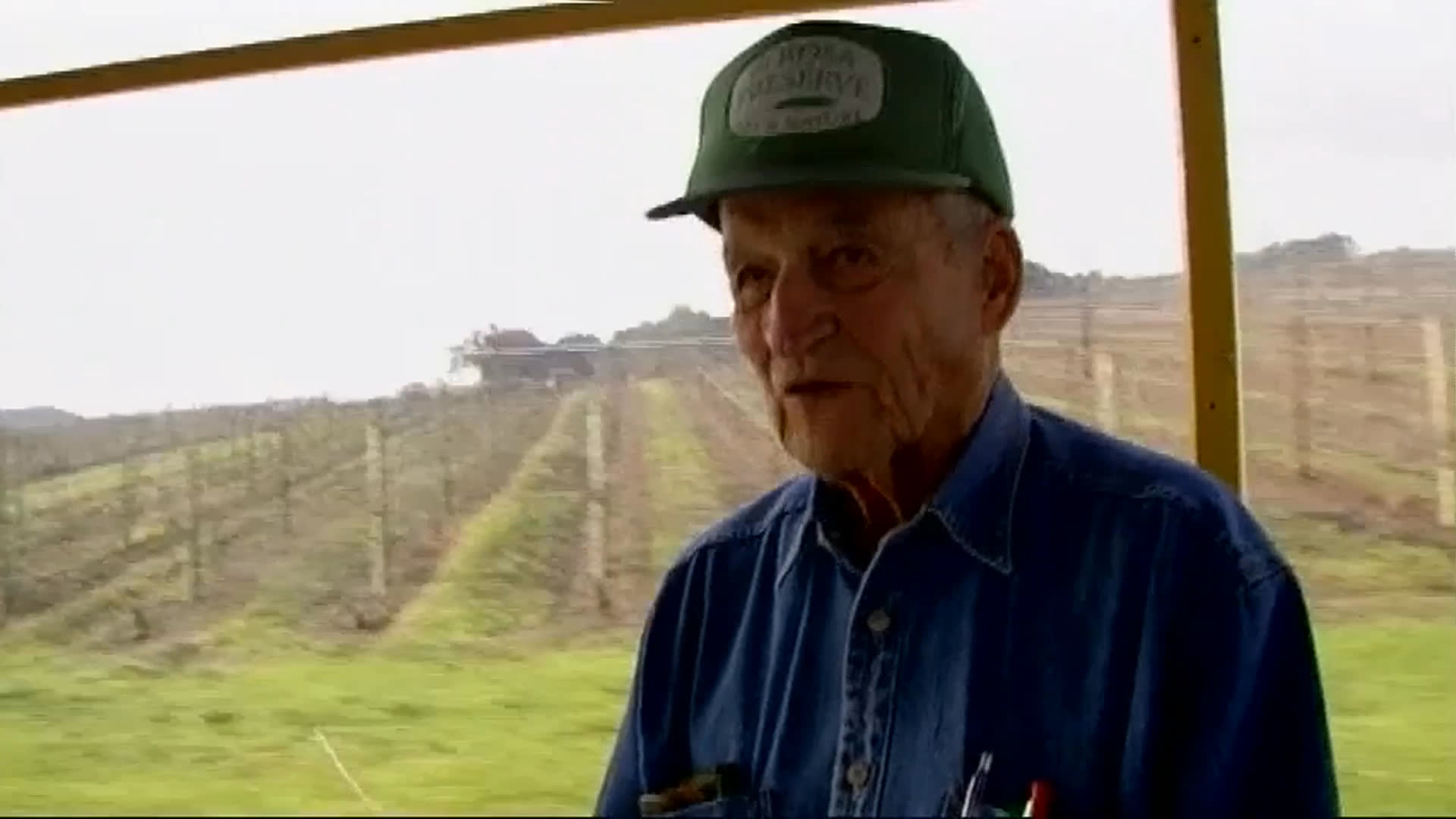An older man in a green cap and work shirt speaks beside vineyard rows, recalling his former work as a reporter for the San Francisco Chronicle. Views of a large agricultural facility and a red pickup near the vineyard provide context.