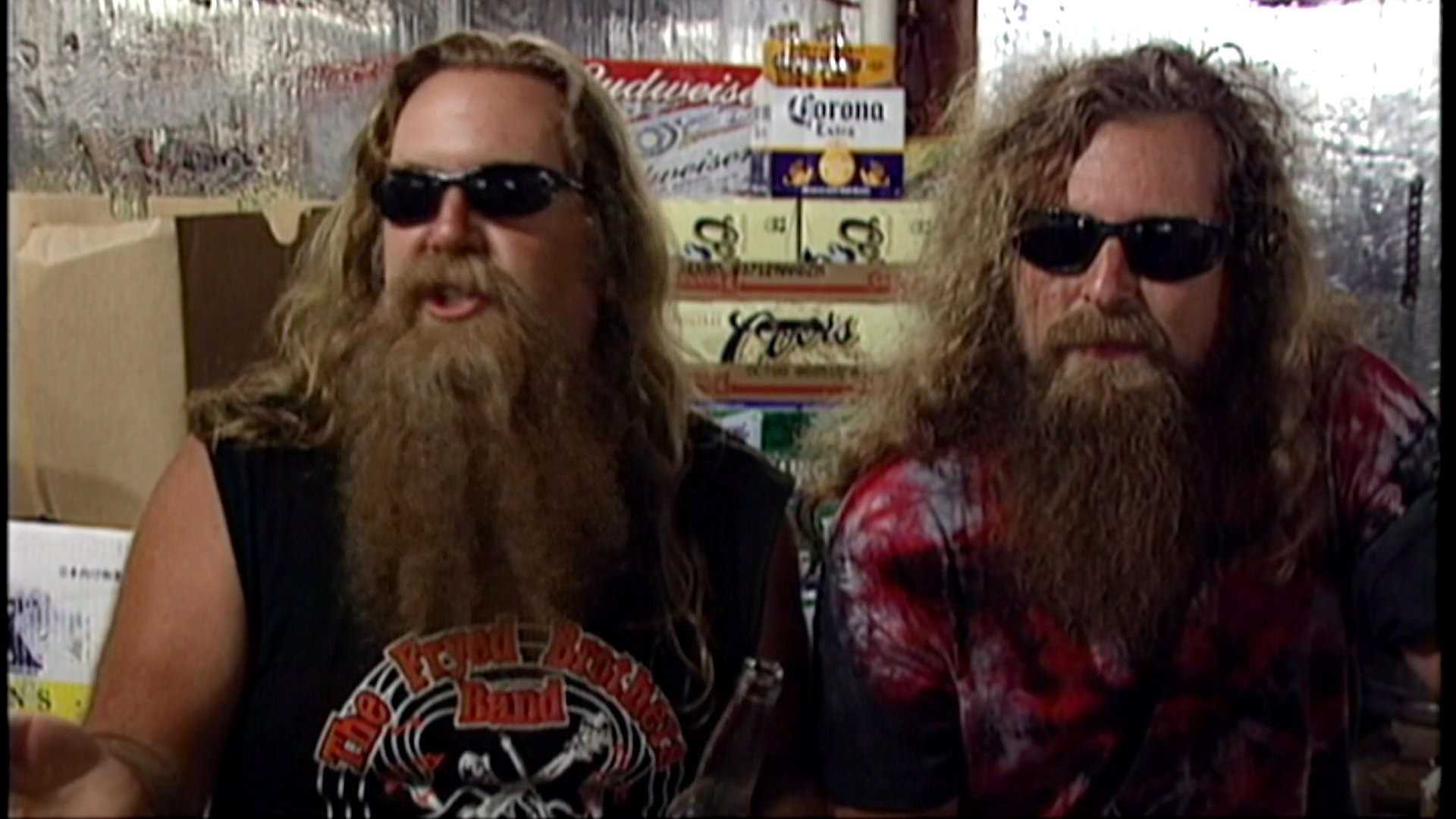 Two long-haired men with thick beards and sunglasses engage in a lively discussion while sitting together in a casual indoor setting.
