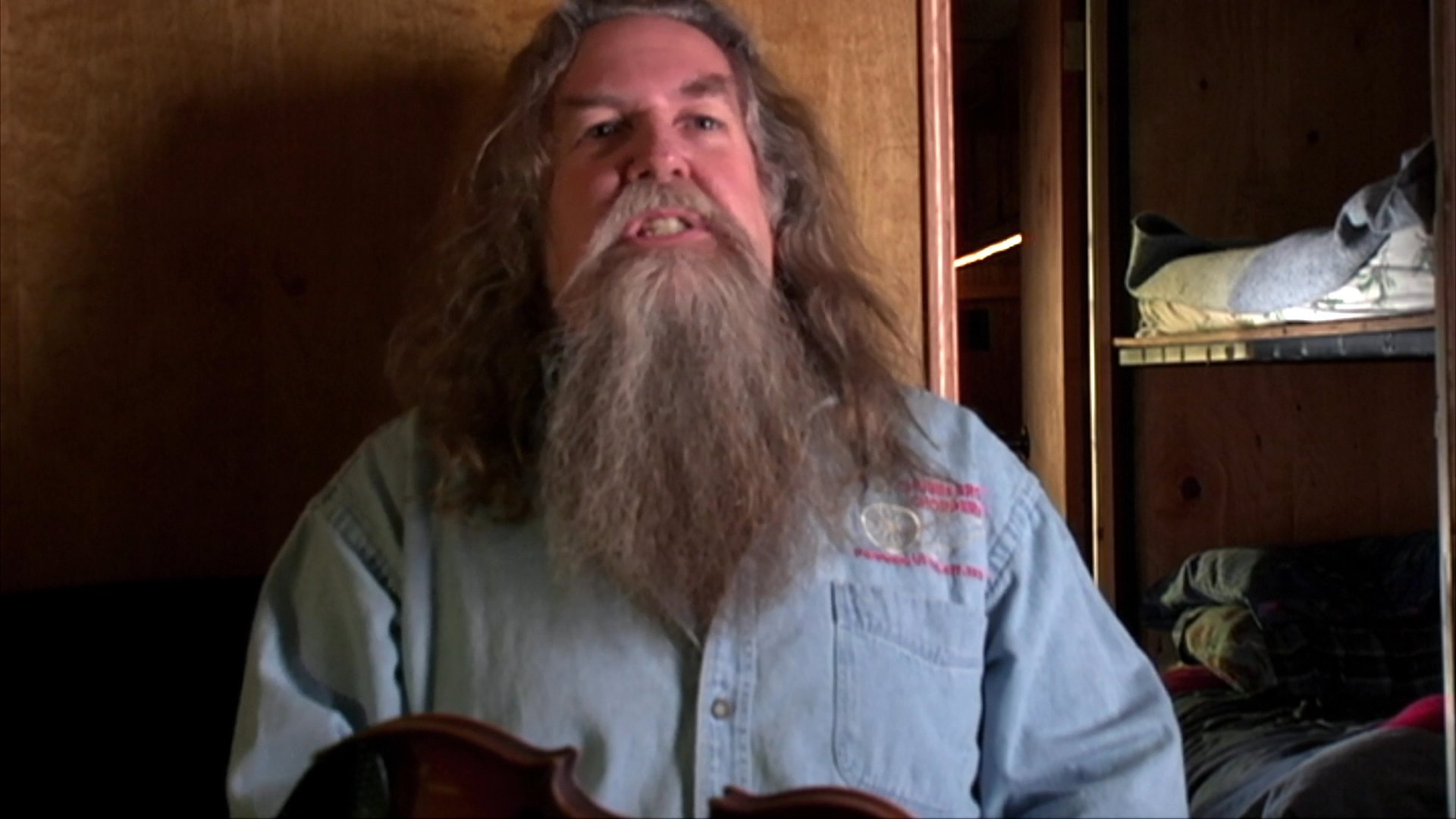 A man with a long beard and long hair speaks expressively while gesturing in an indoor setting with wood paneling. He conveys a narrative through a series of animated facial expressions and hand motions.