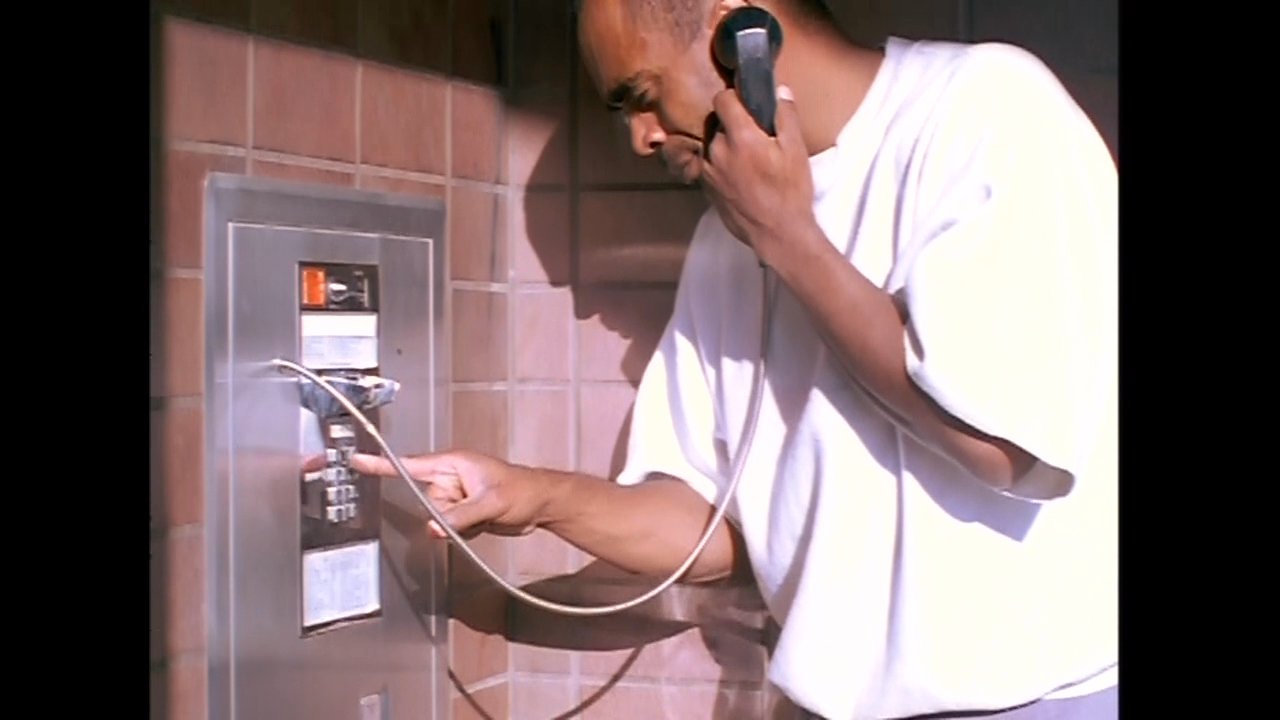 A man makes a tense telephone call from a wall-mounted payphone before shifting to a scene of two men sitting together in a room. The sequence highlights themes of isolation and communication within a confined environment.